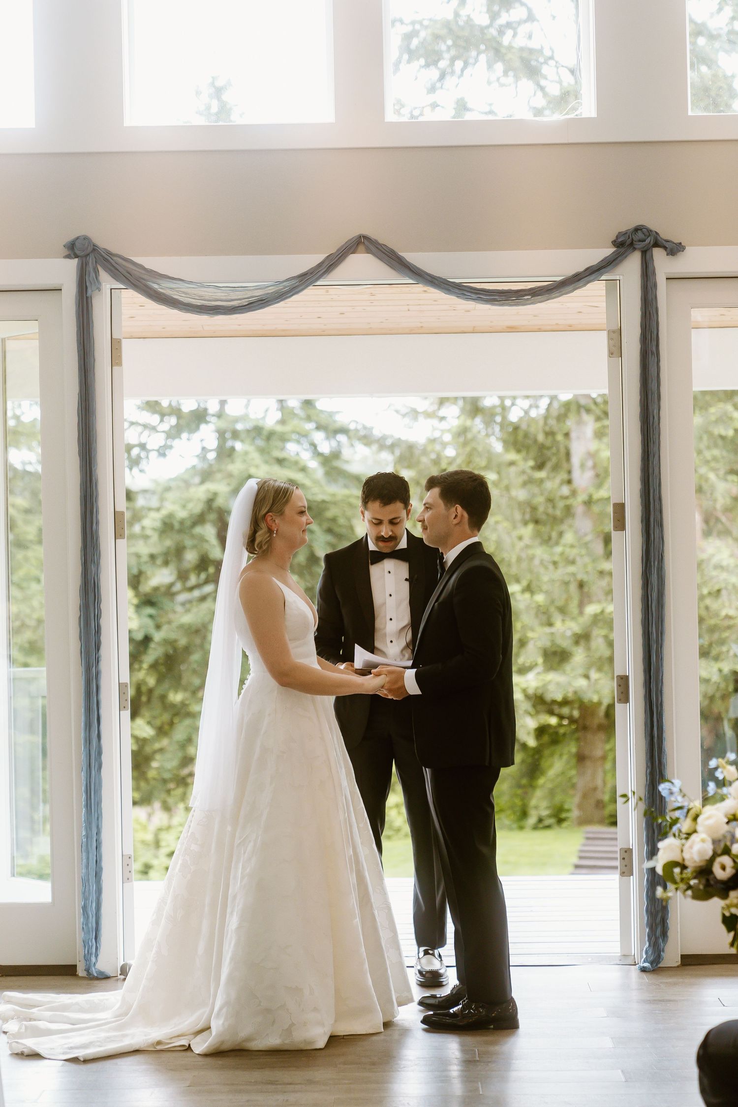 Bride and groom hold hands during ring exchange with smiling officiant at indoor wedding ceremony.
