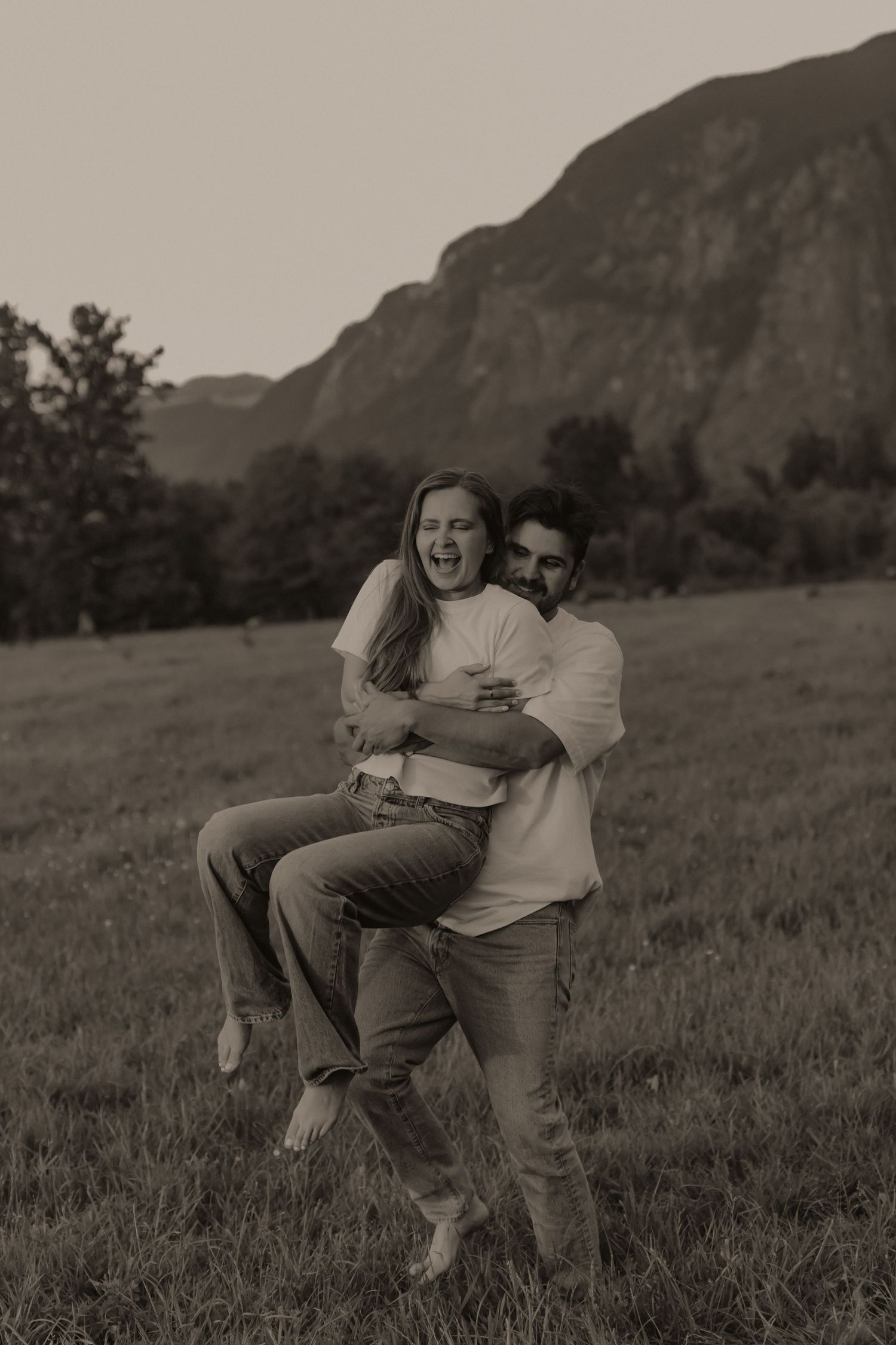 Couple laughing and playing in a mountain meadow, man lifting woman in a joyful embrace, sepia tone.