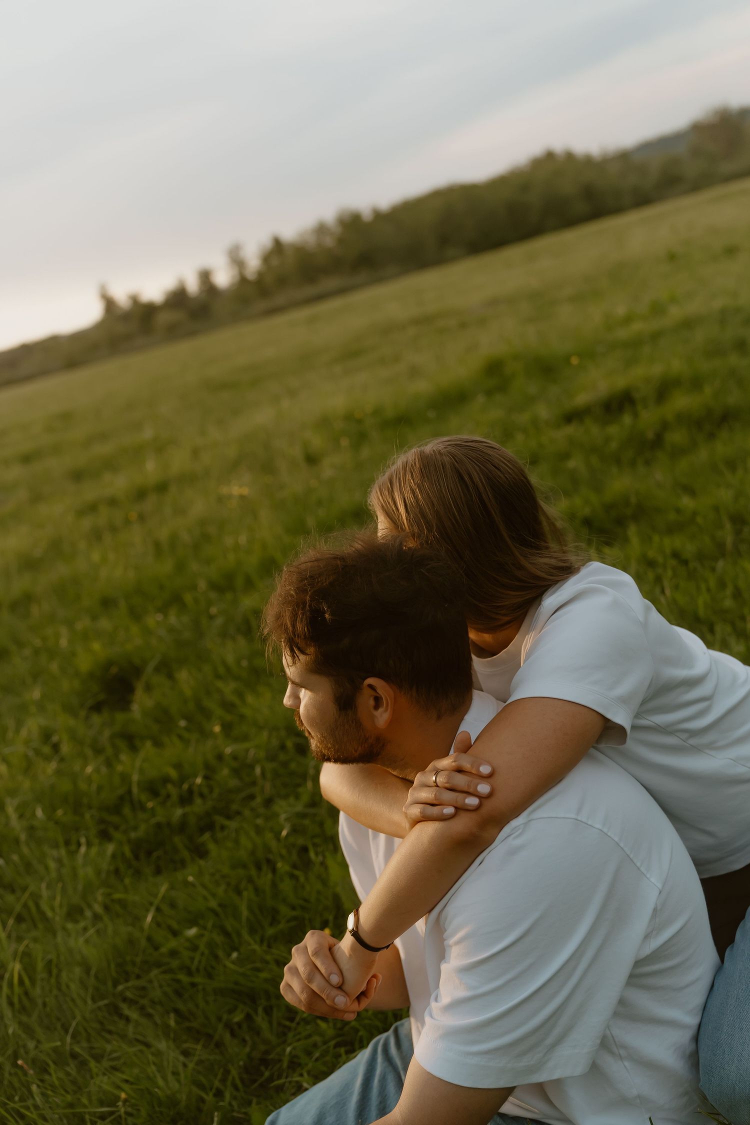 Couple sitting on green grass field, woman hugging man from behind while holding hands at sunset.