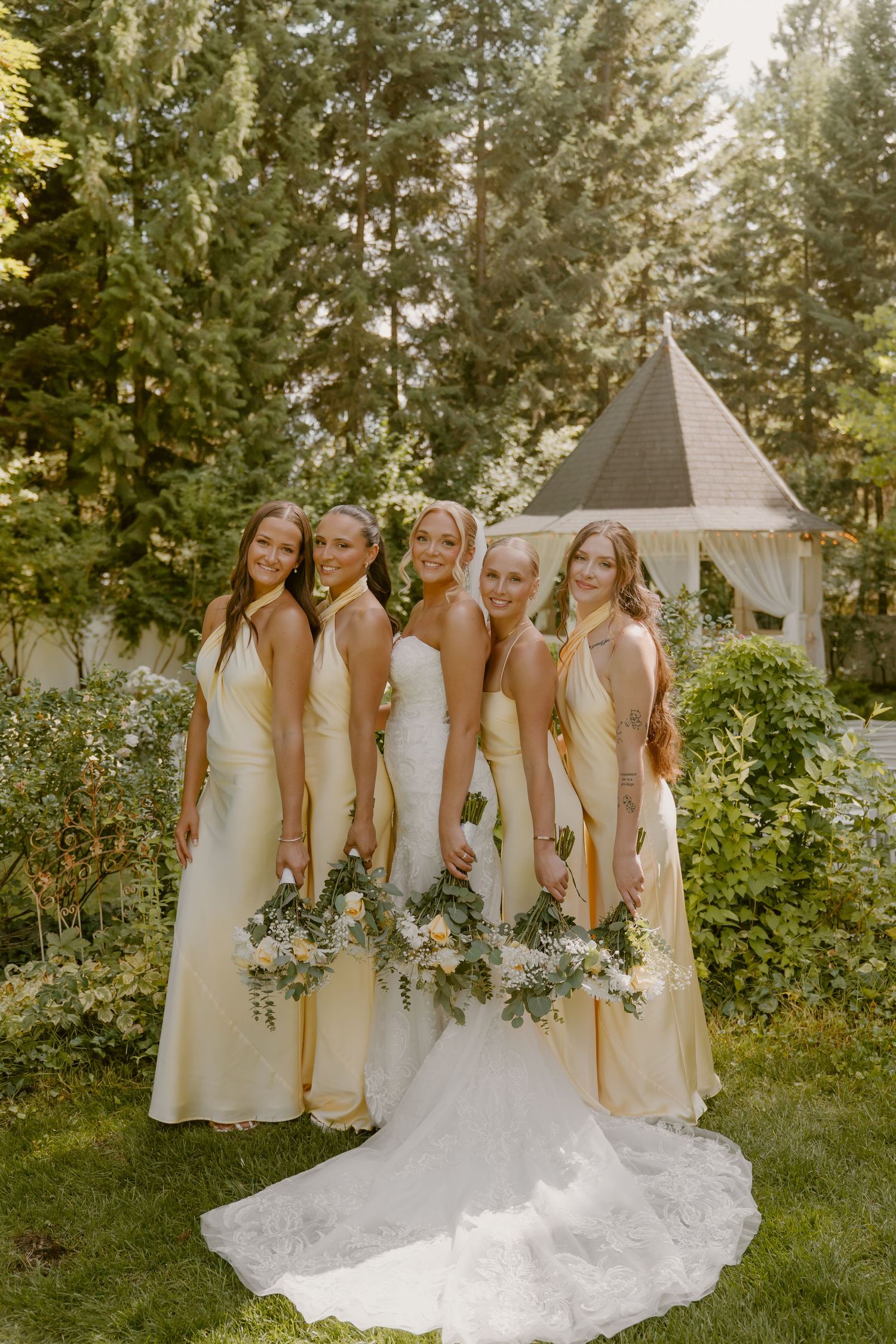 A wedding party in light colored dresses poses together outdoors among evergreen trees and greenery.