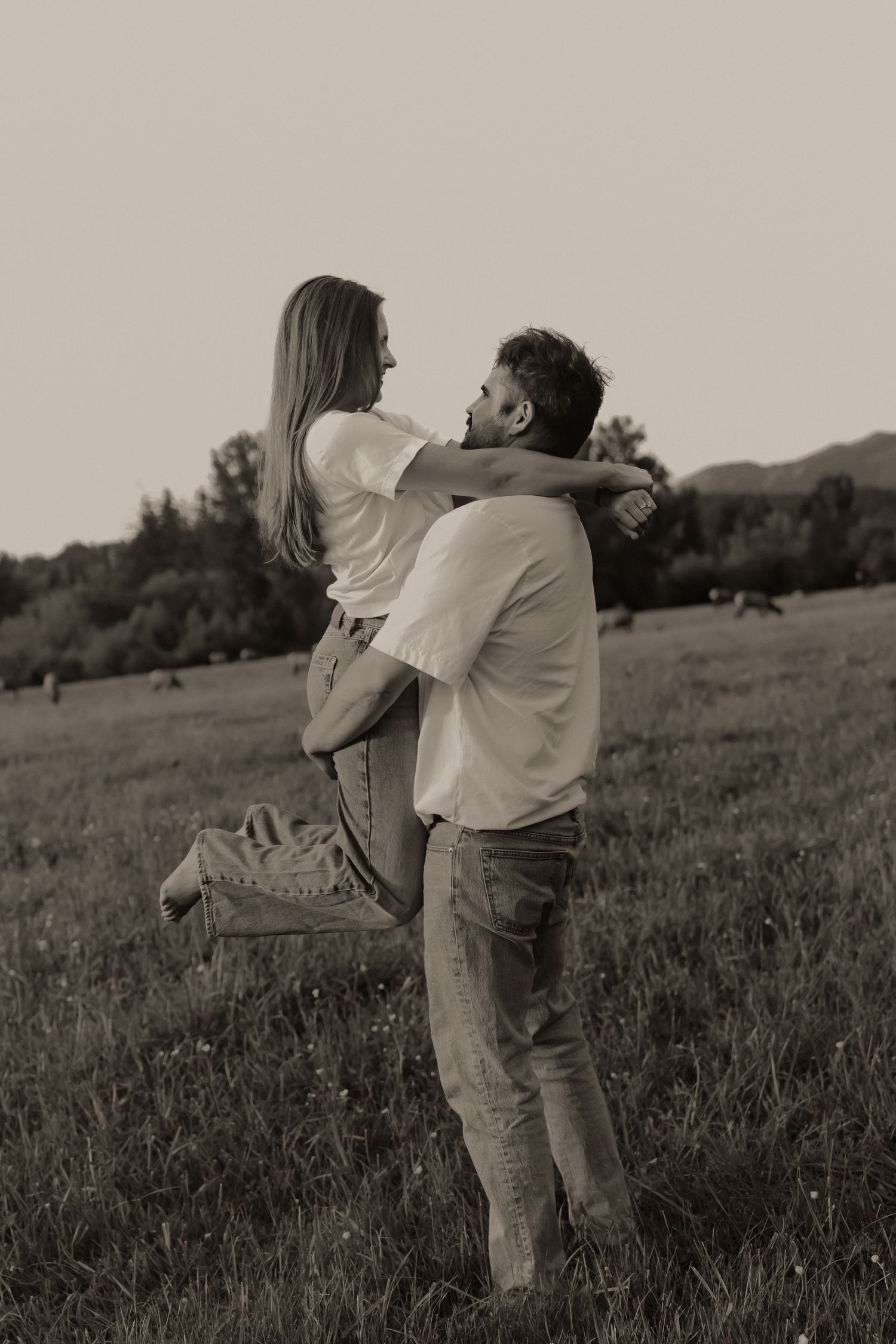 A couple shares a romantic moment in a field, as the man lifts the woman in his arms in a sepia-toned photo.