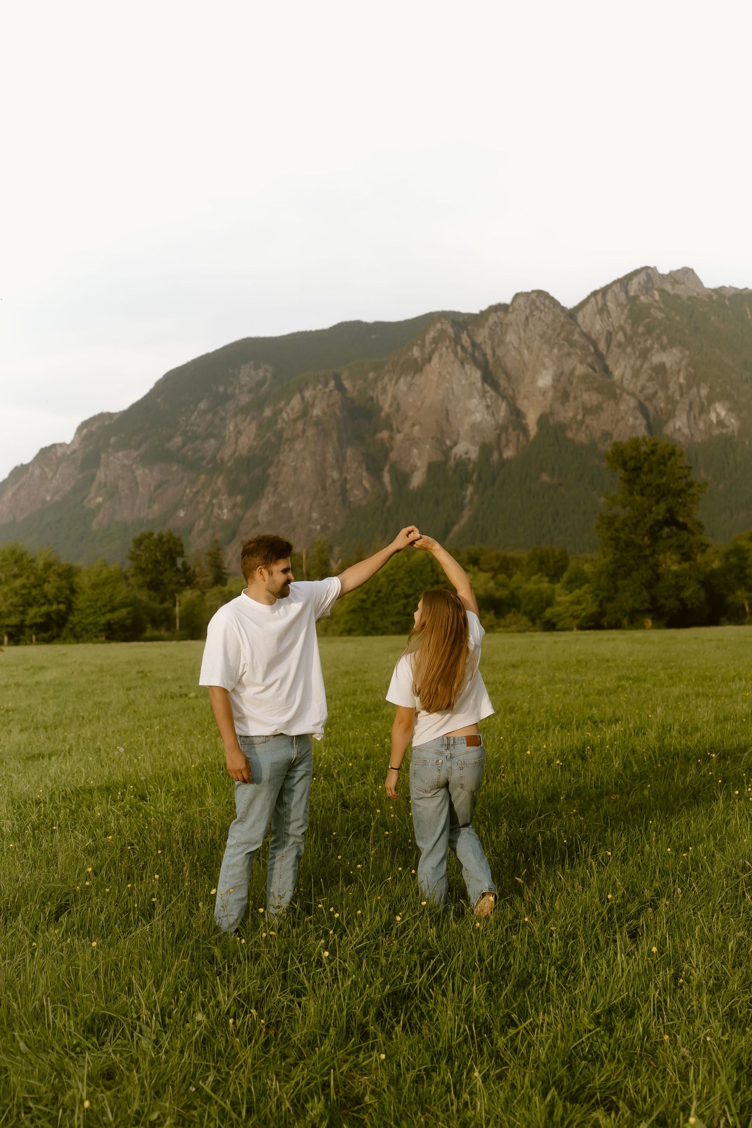 Couple in matching white tees and jeans dancing in a green field with a rocky mountain backdrop.