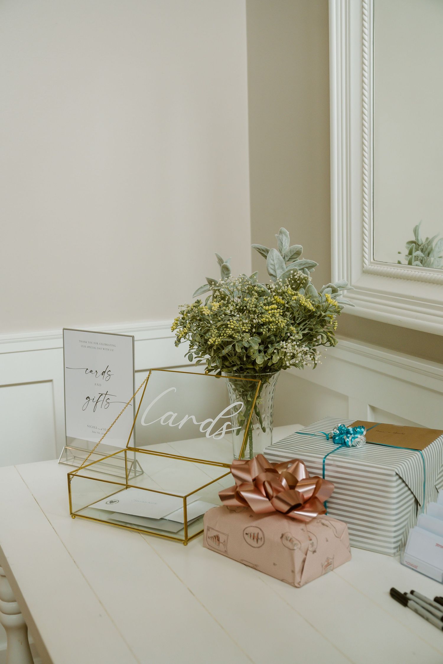 Elegant wedding gift table with wrapped presents, gold card box, greenery bouquet, and acrylic cards sign on white table.