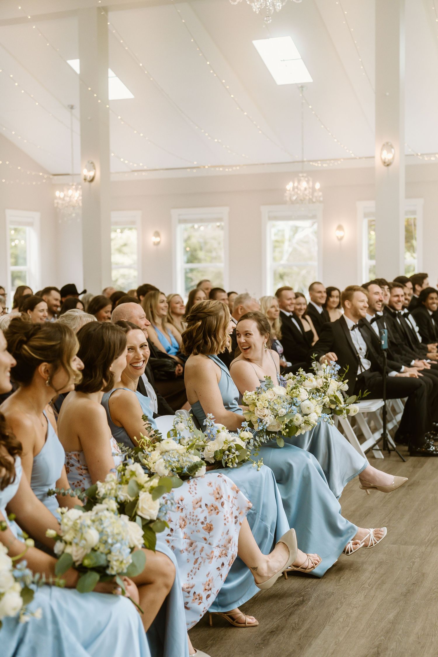 Bridesmaids in blue dresses hold white floral bouquets while seated at a bright wedding ceremony.