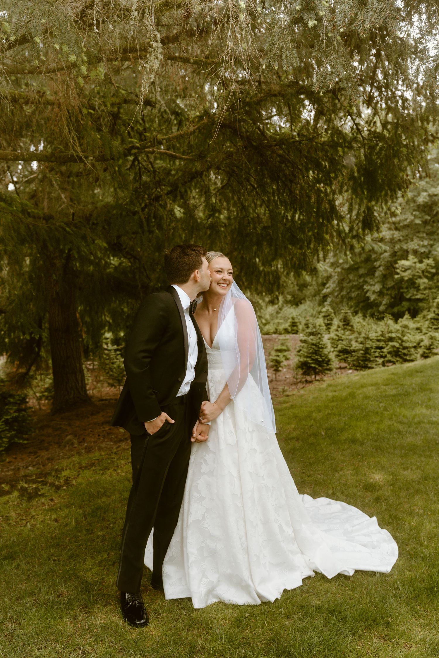 Bride and groom share a romantic moment outdoors, smiling under lush green trees.