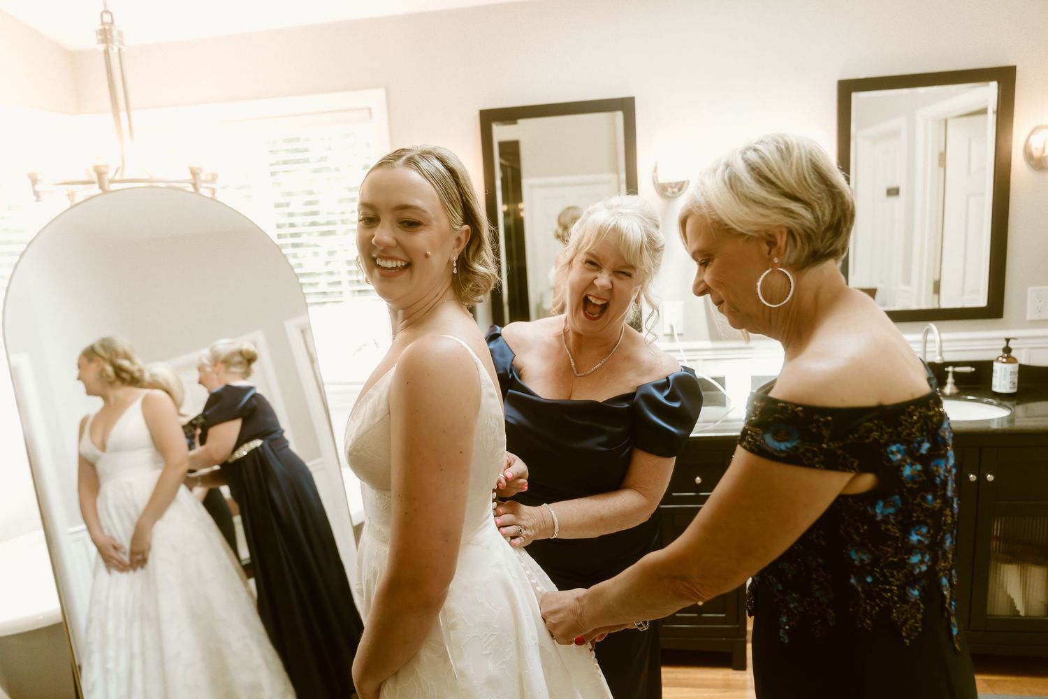 Bride laughing in white wedding gown with two women helping her dress in a bright bridal suite mirror.