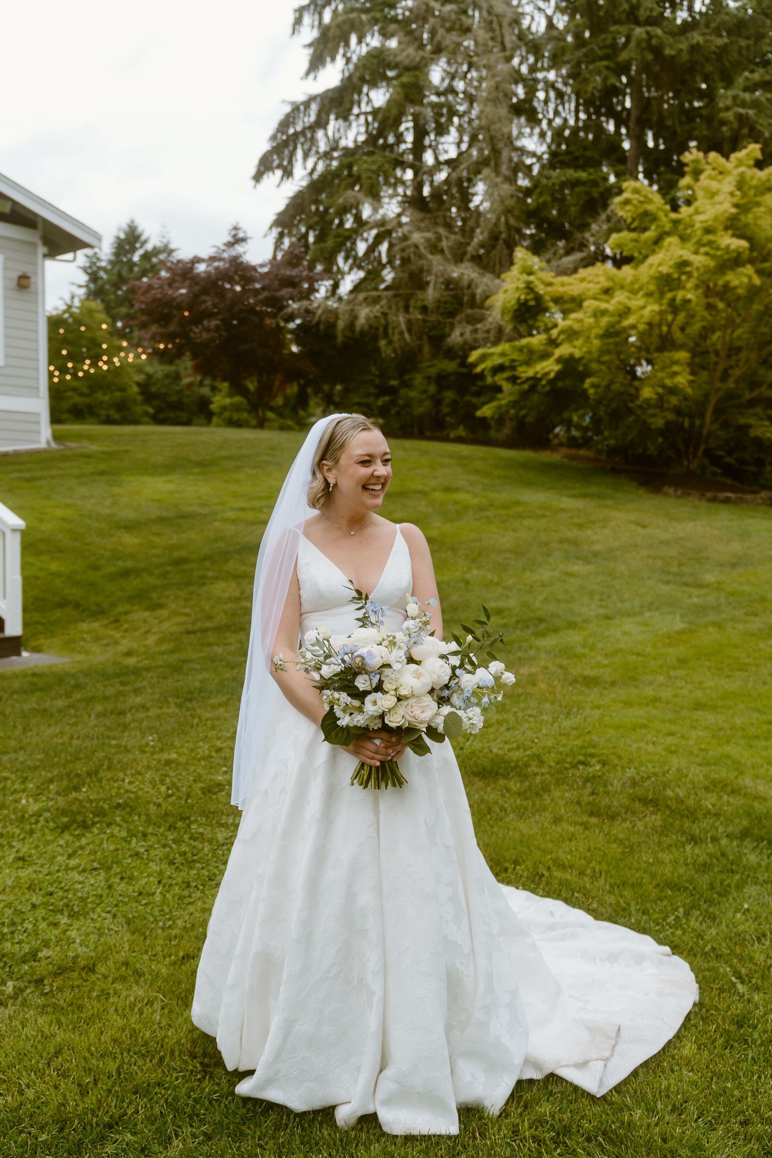 Smiling bride in white ball gown with veil holds white bouquet on green lawn with trees and string lights.