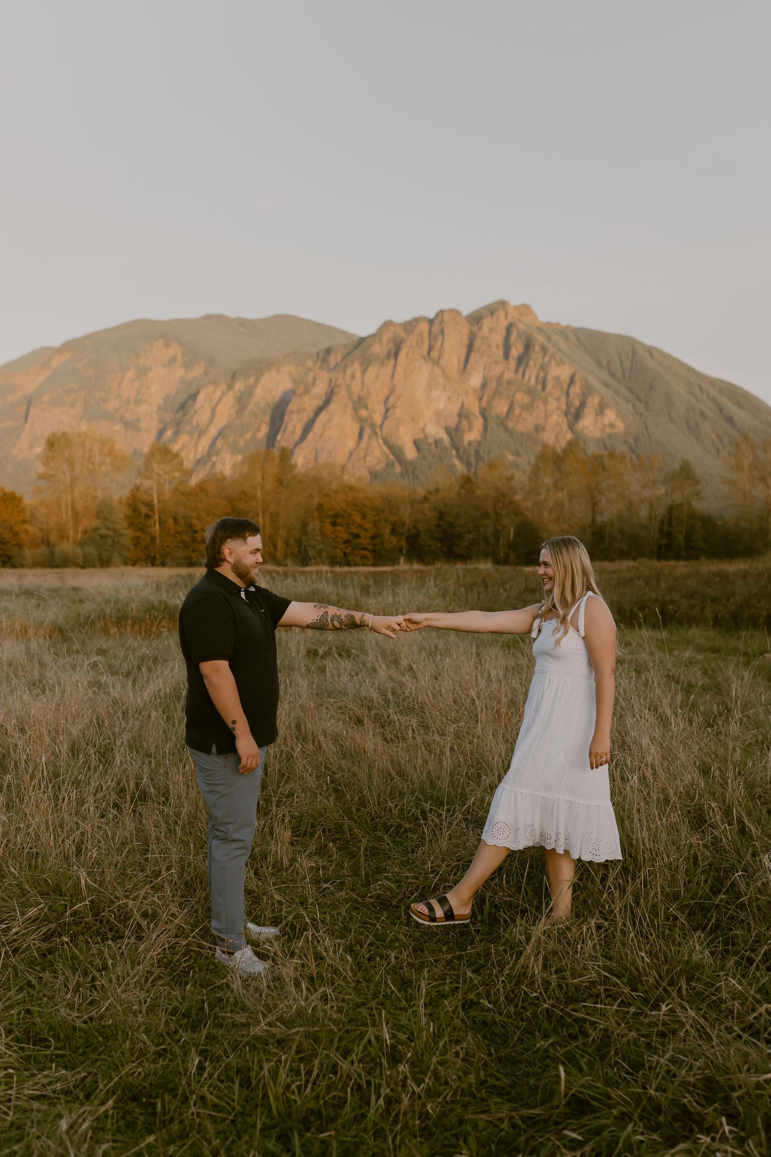 Couple holding hands and dancing in a golden grassy field at sunset with a rugged mountain and autumn trees in the background.