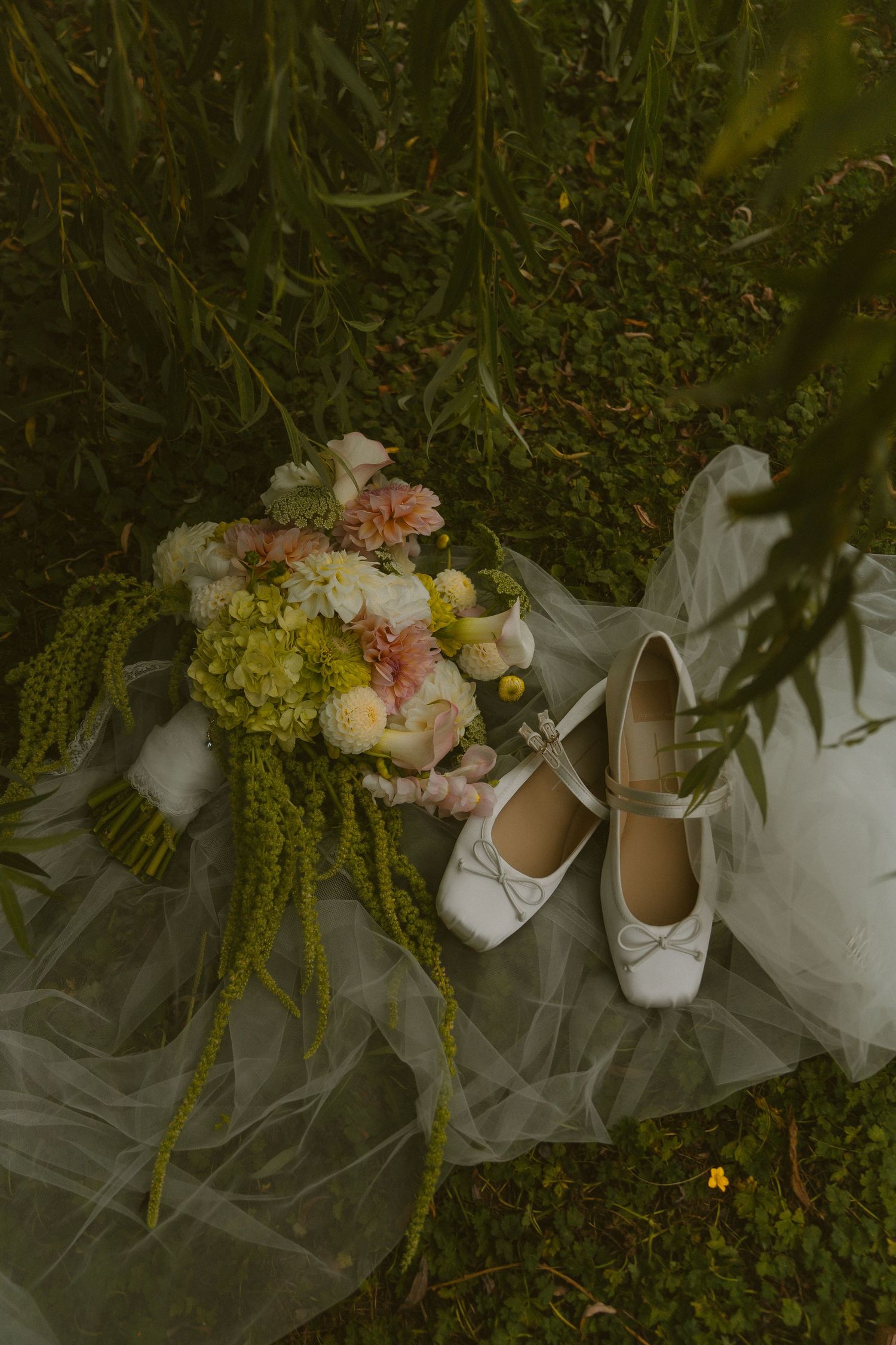 Bridal flat shoes with bow, white veil, and pink floral bouquet with greenery arranged on lush grass.