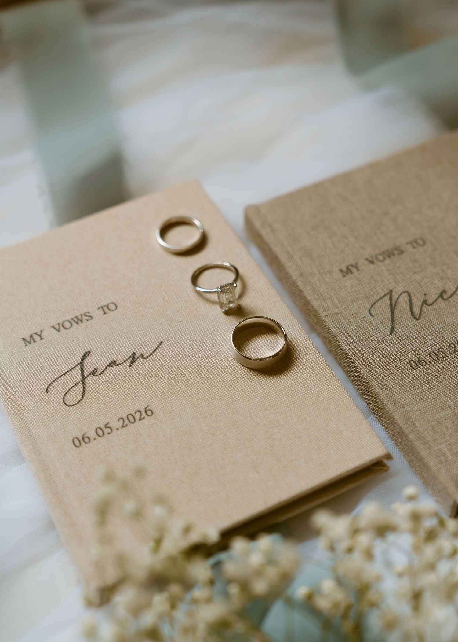 Two personalized wedding vow books with gold rings resting on top, dated 06.05.2026, surrounded by delicate white flowers.