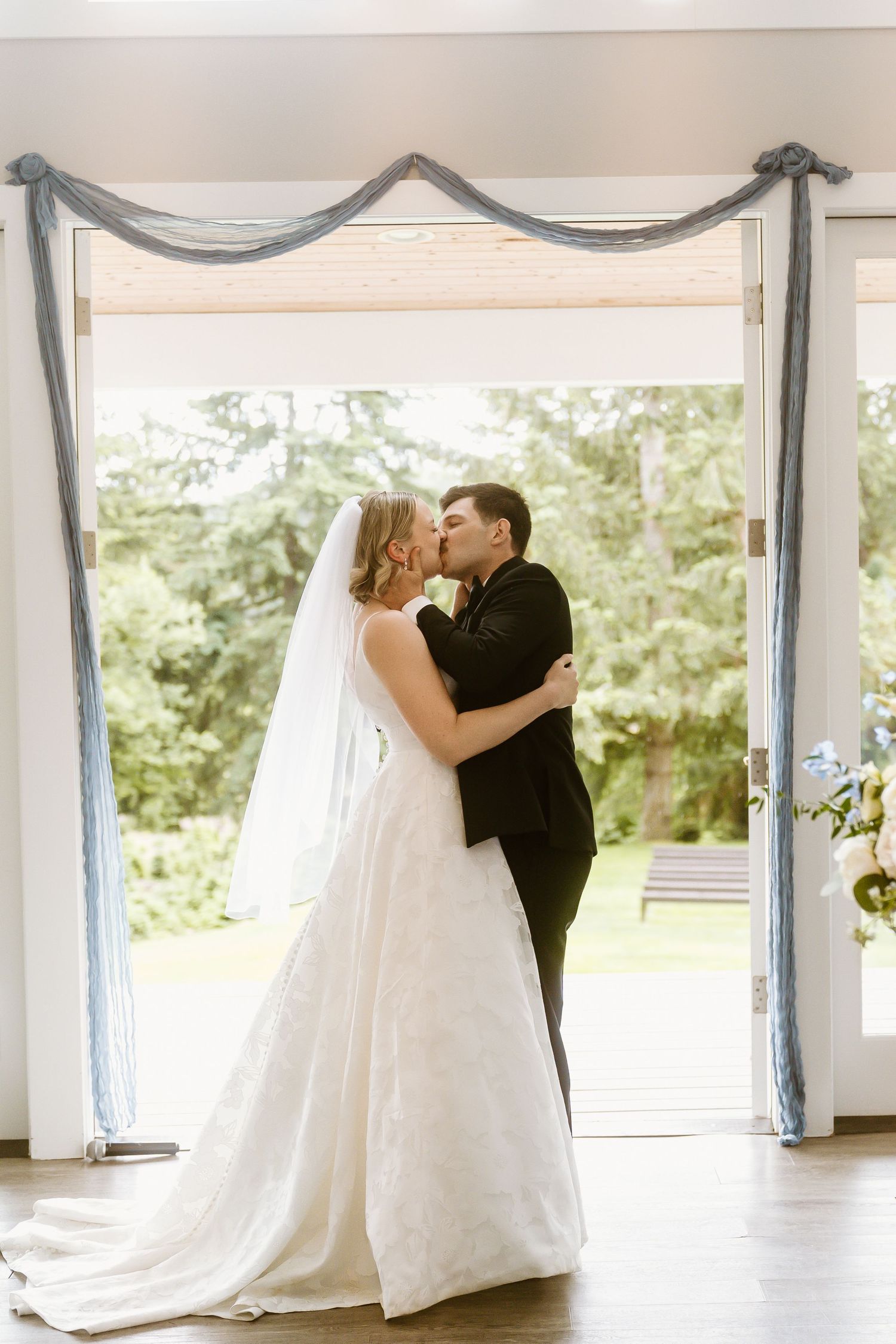 Bride and groom share a romantic first kiss framed by blue draped doors at their wedding.