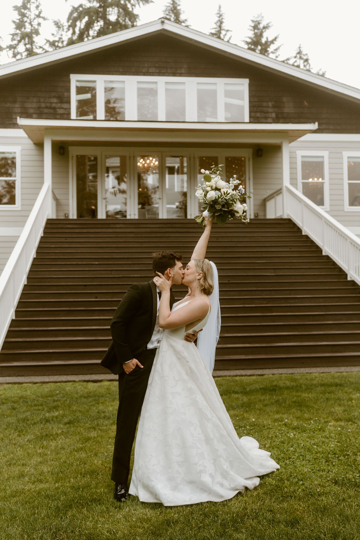 Bride raises bouquet while kissing groom in front of elegant venue staircase on their wedding day.