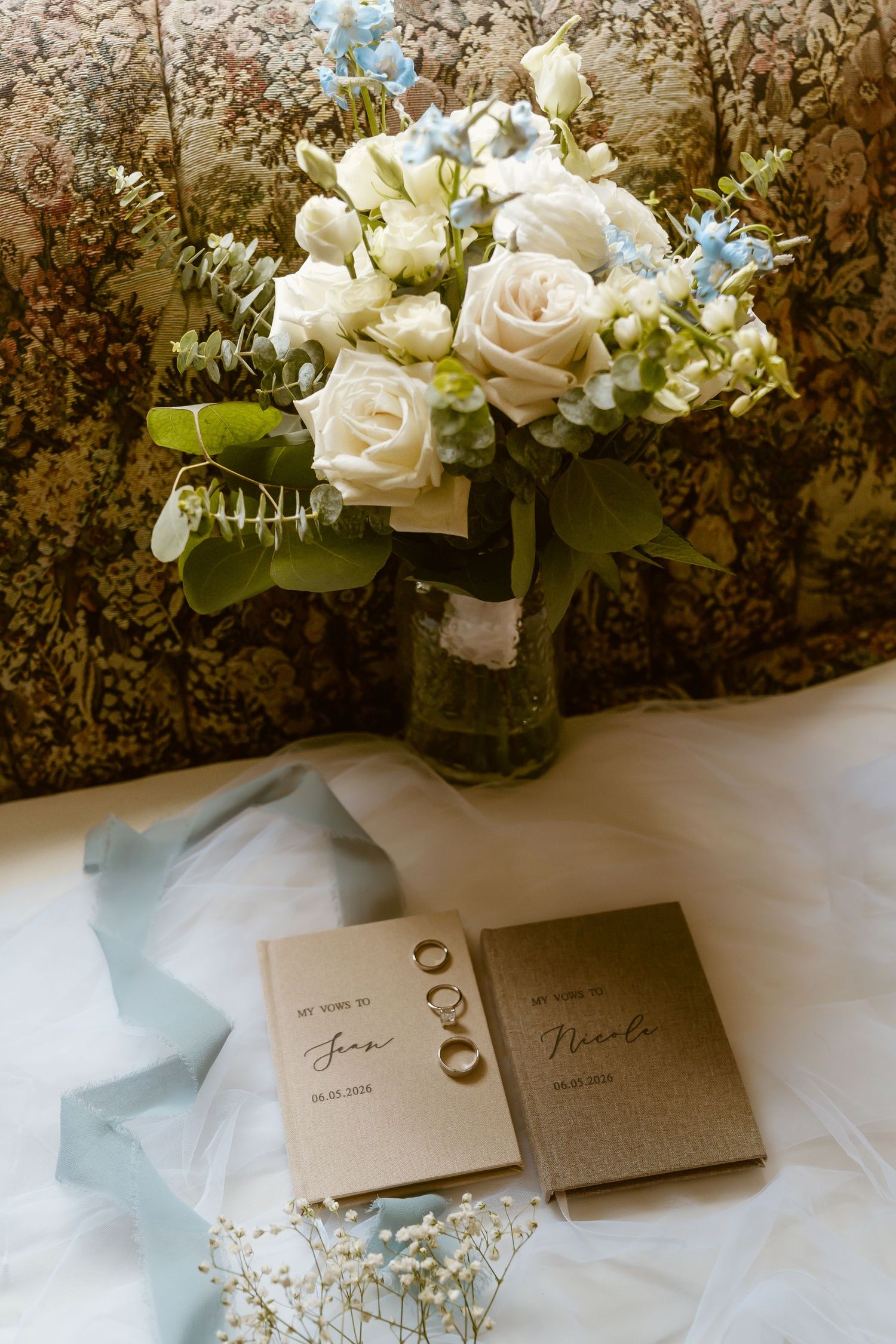 Wedding flat lay with white roses, blue florals, vow books, and gold rings on white tulle fabric.