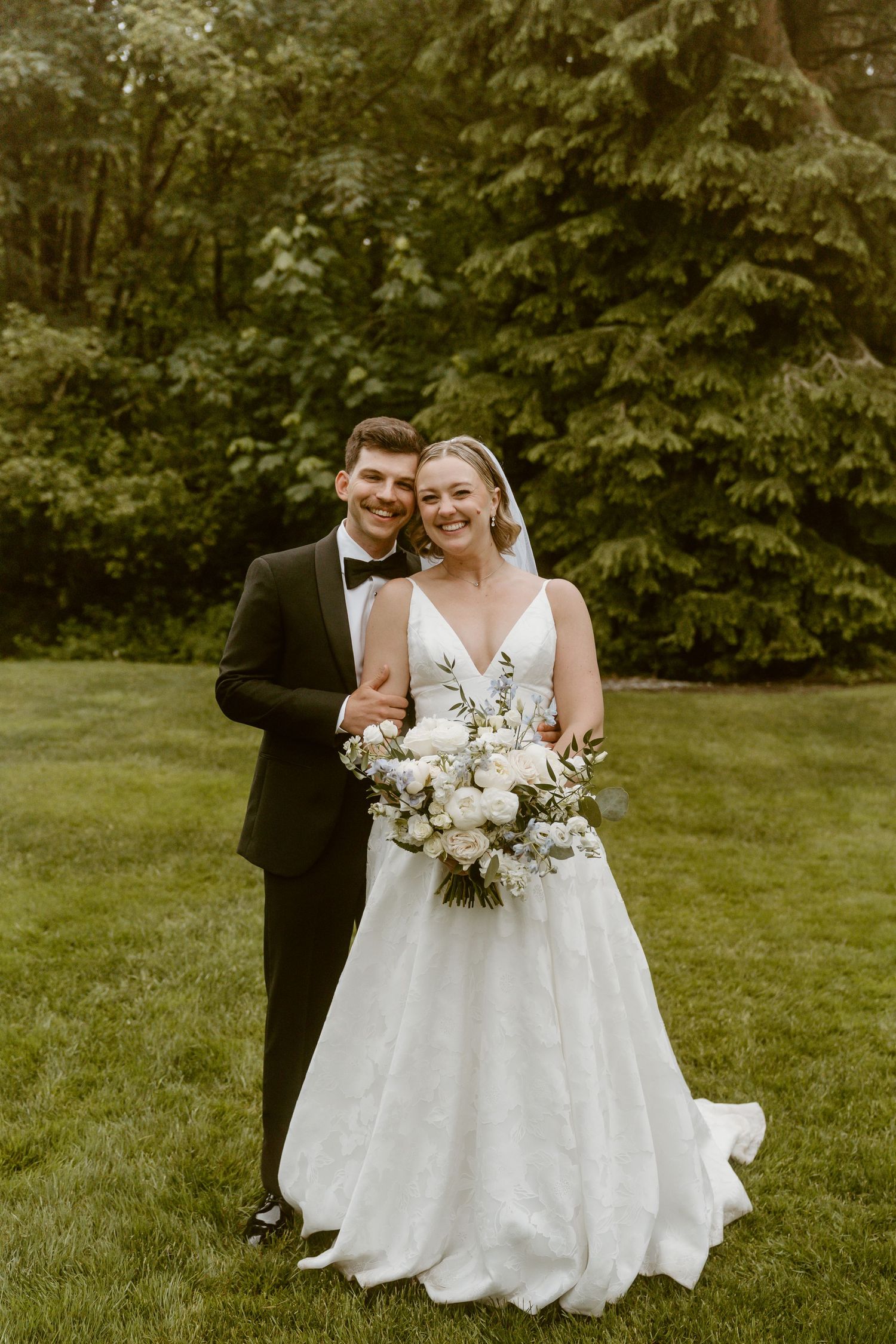 Smiling bride holding white floral bouquet stands with groom in black tuxedo on green lawn.