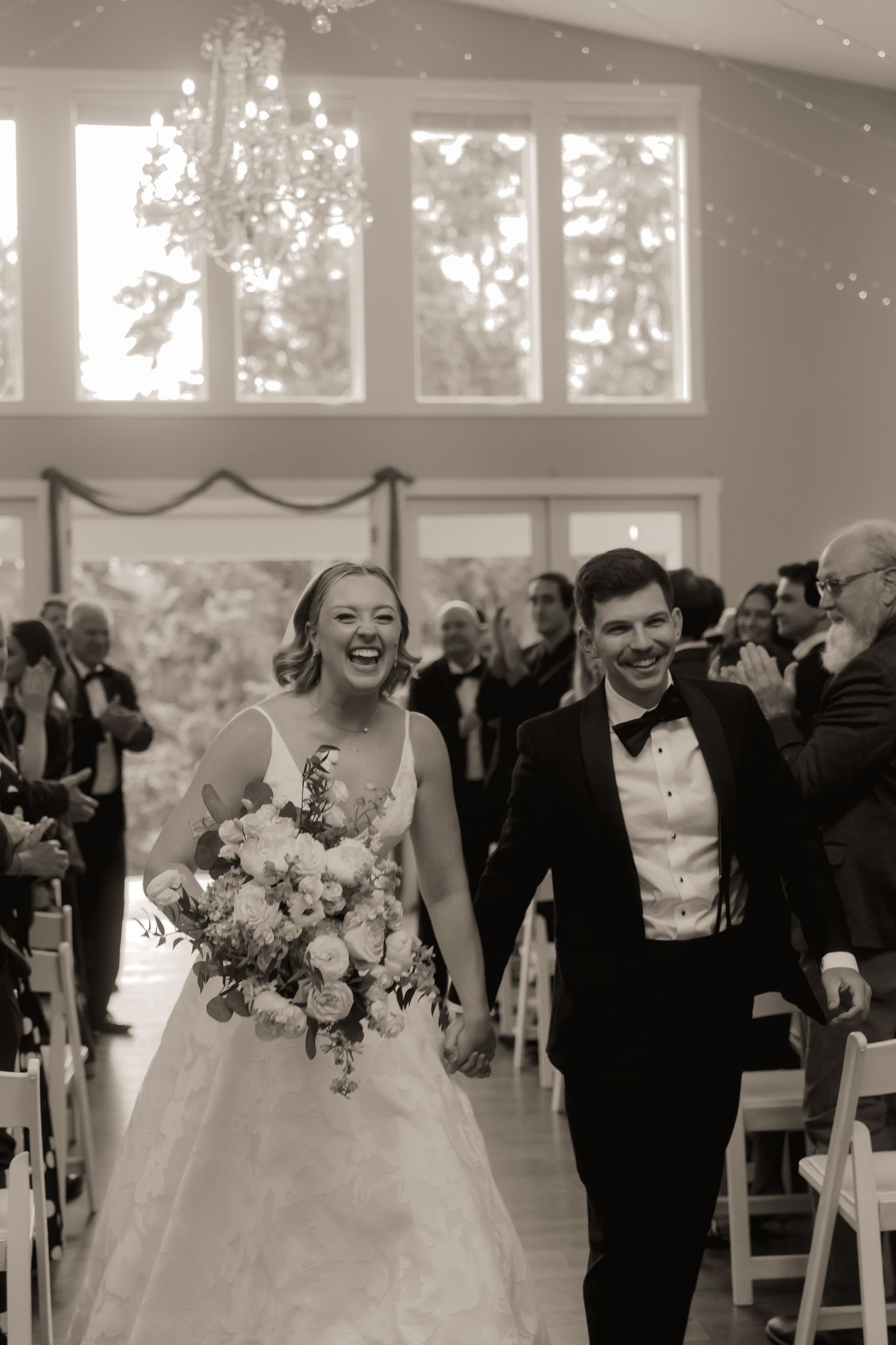 Black and white photo of smiling newlyweds holding hands walking down wedding ceremony aisle.