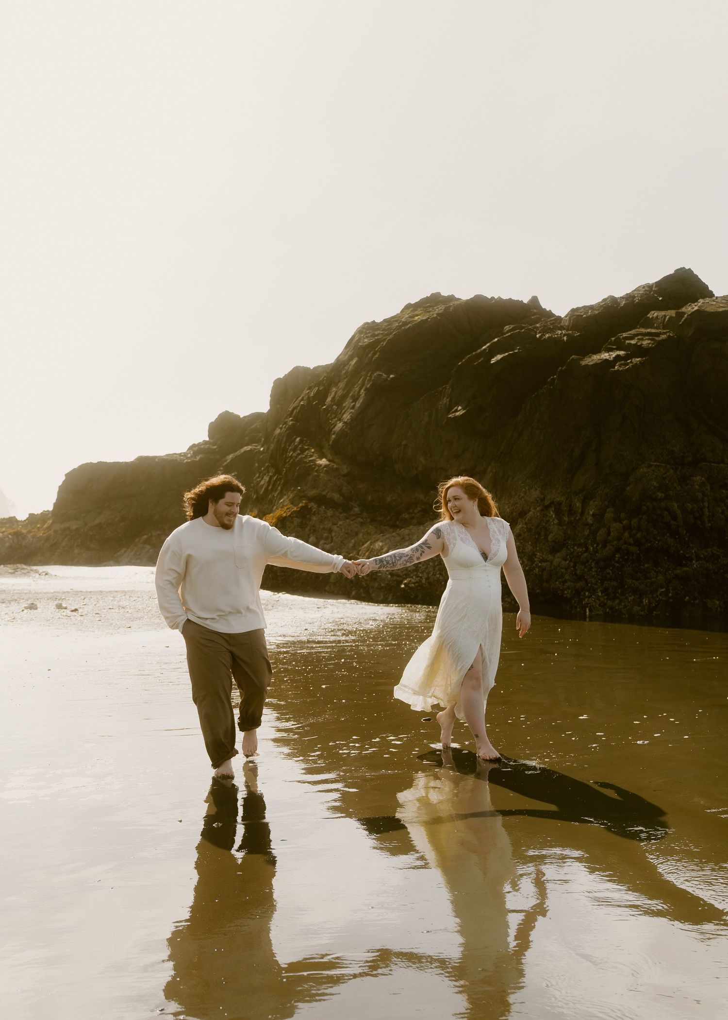Couple holding hands walking on a reflective beach with rocky cliffs, woman in white dress, moody coastal elopement photo.