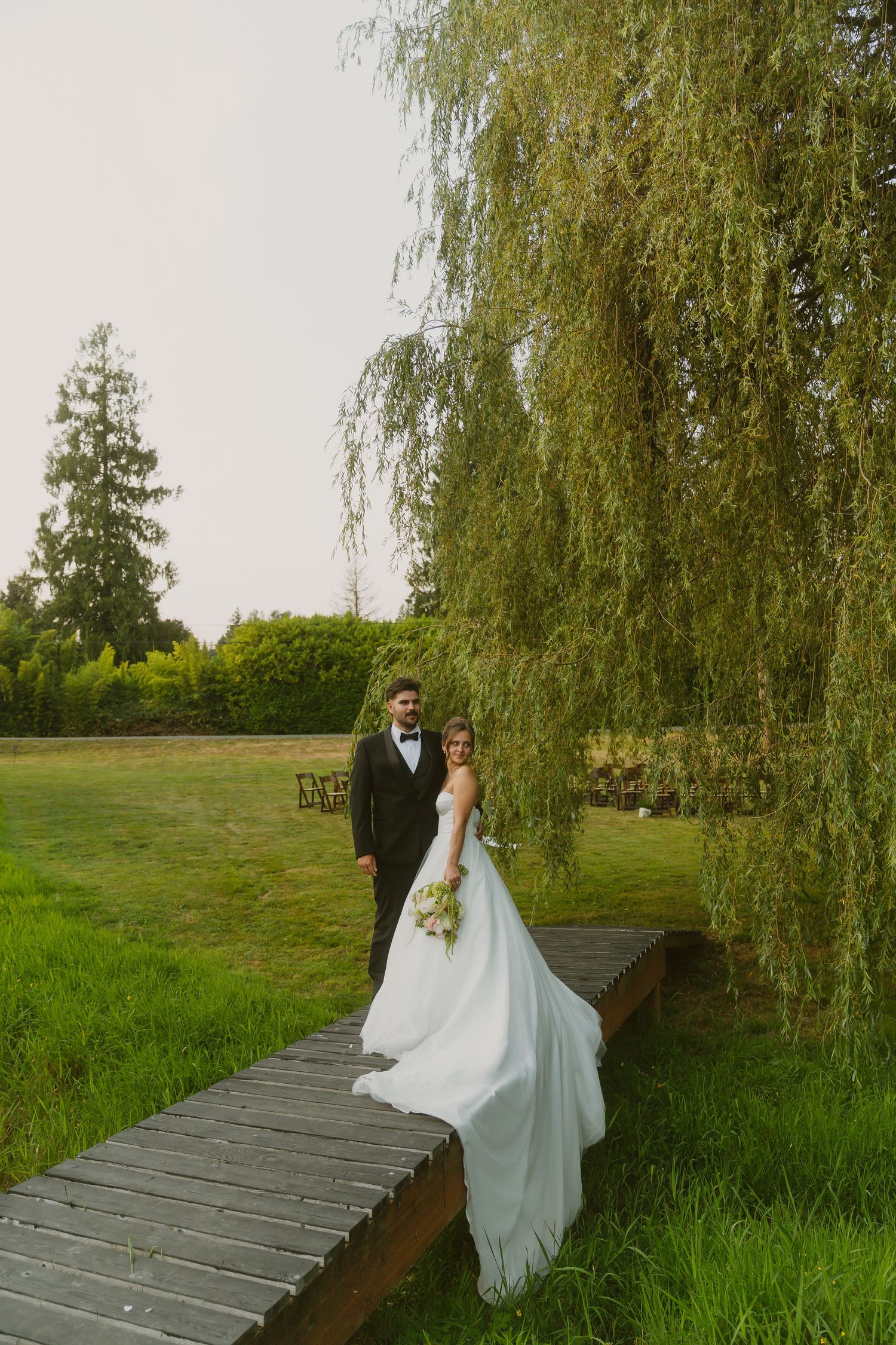 Bride and groom pose on a wooden bridge beneath a weeping willow tree in a lush green garden setting.