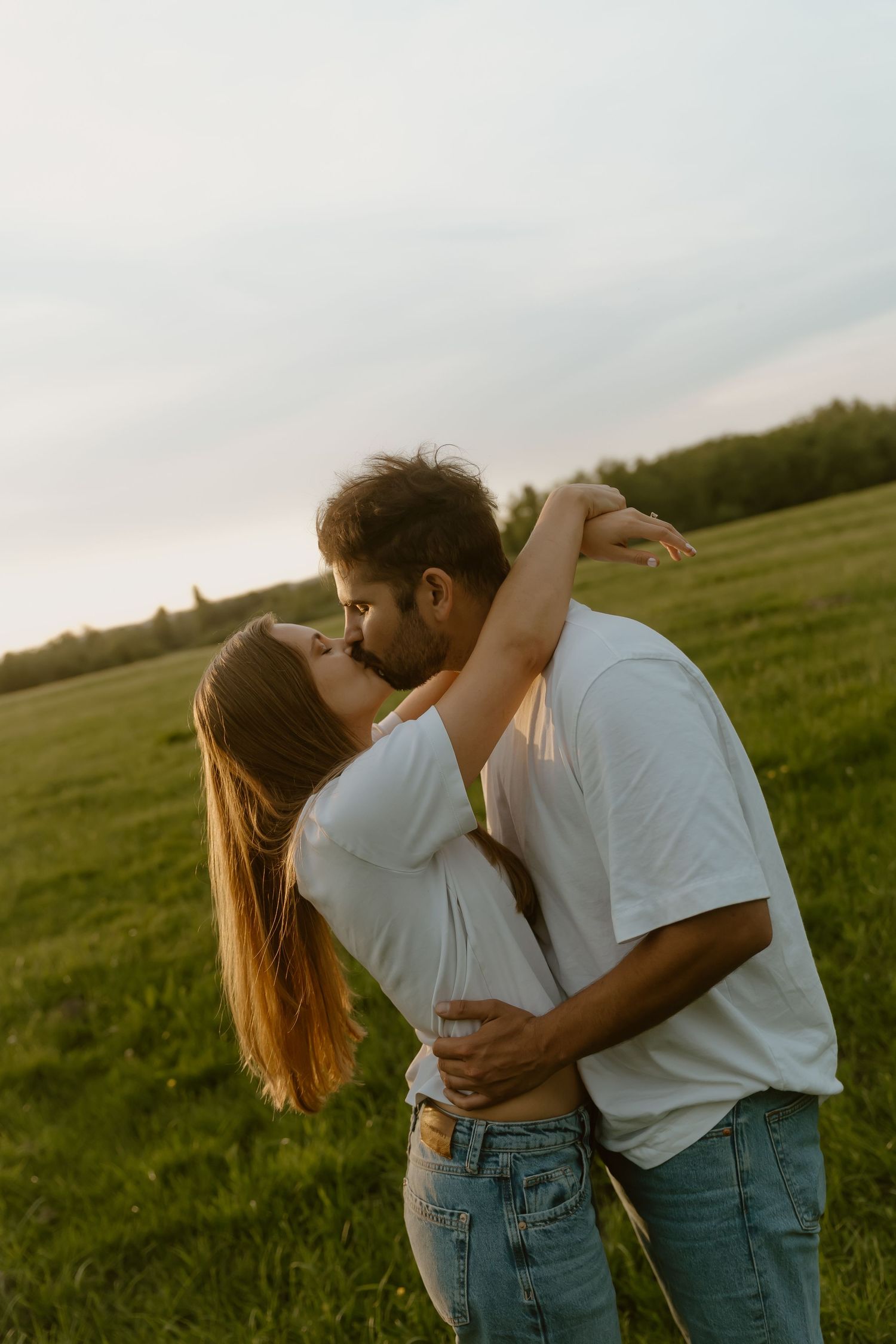 Couple sharing a romantic kiss in a green field at sunset, both wearing white shirts and jeans.