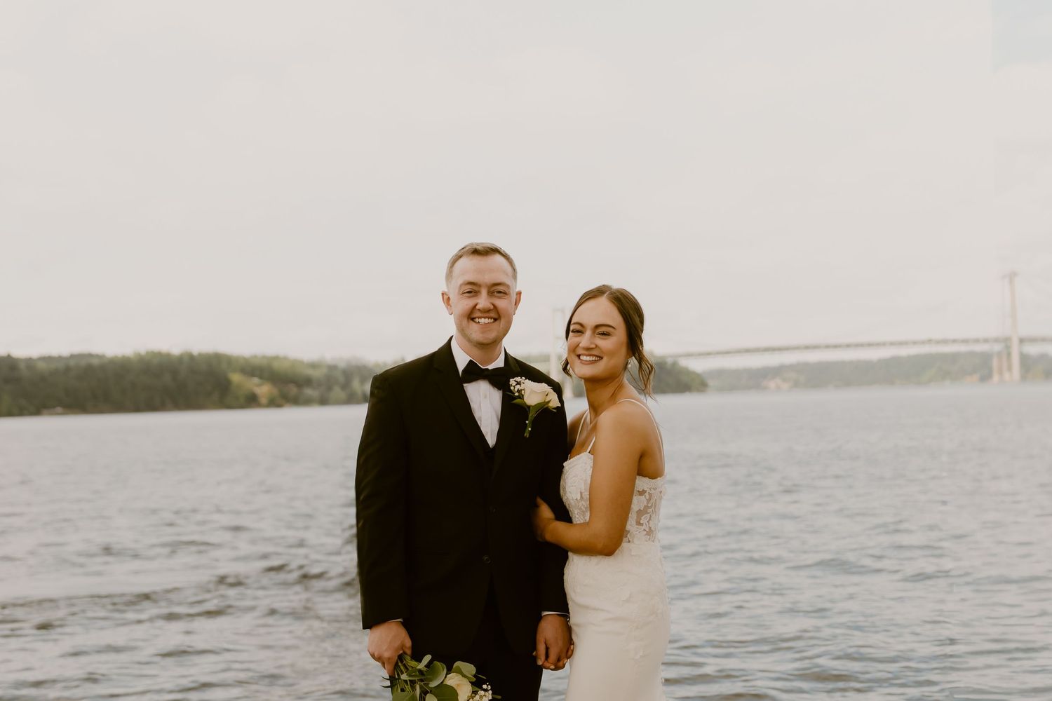 Bride and groom smiling together by waterfront with suspension bridge in background at outdoor wedding.