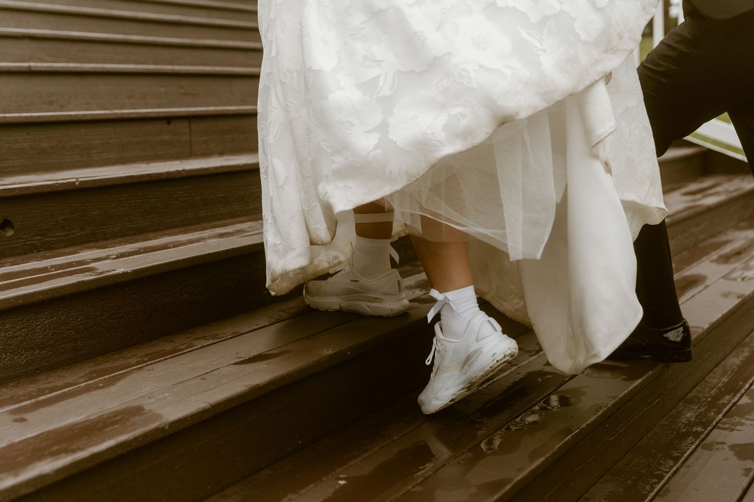 Bride in white wedding dress wearing white sneakers walking up wooden steps with groom in black dress shoes.