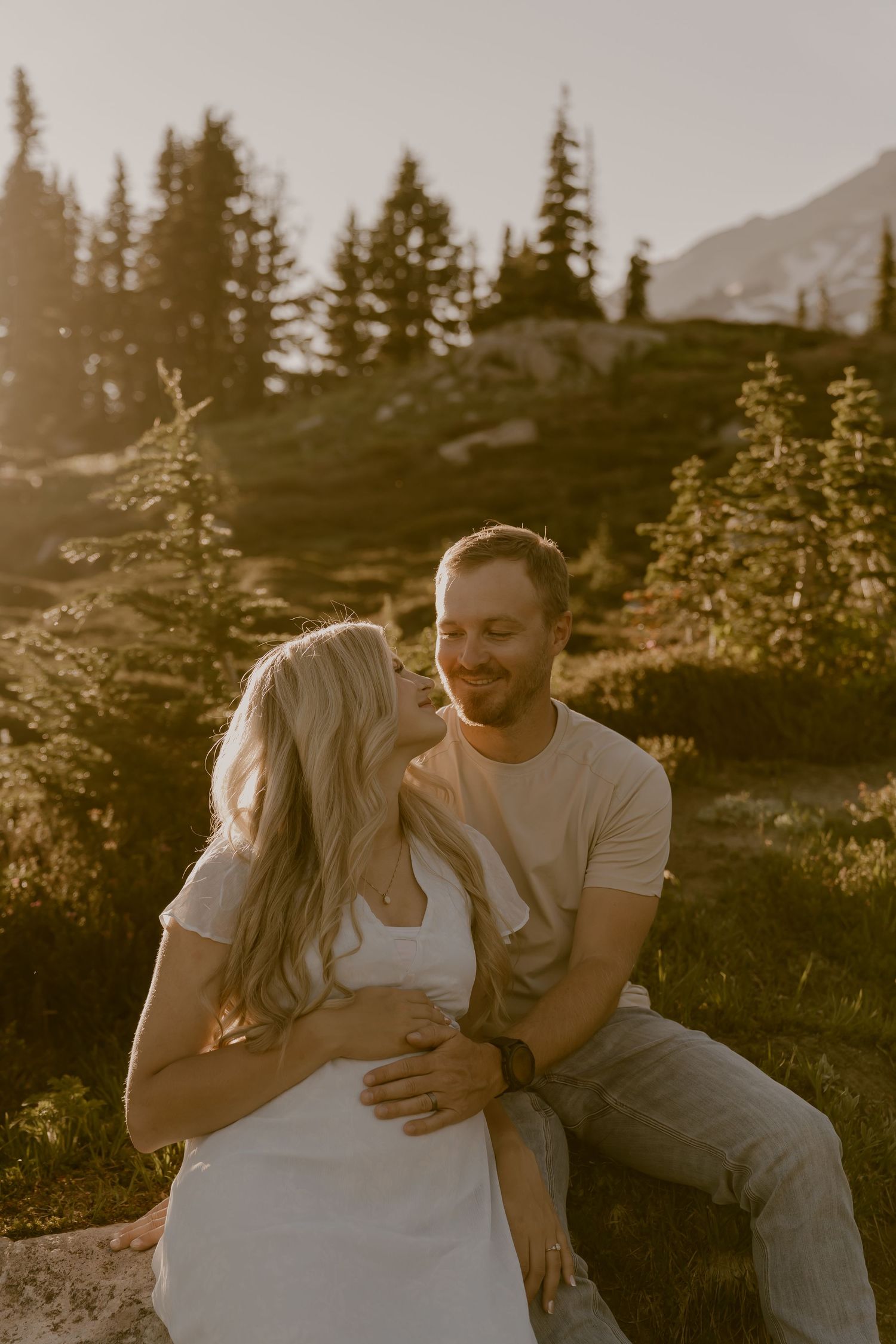 Couple sitting together in a sunlit mountain meadow surrounded by evergreen trees, sharing a tender moment at golden hour.