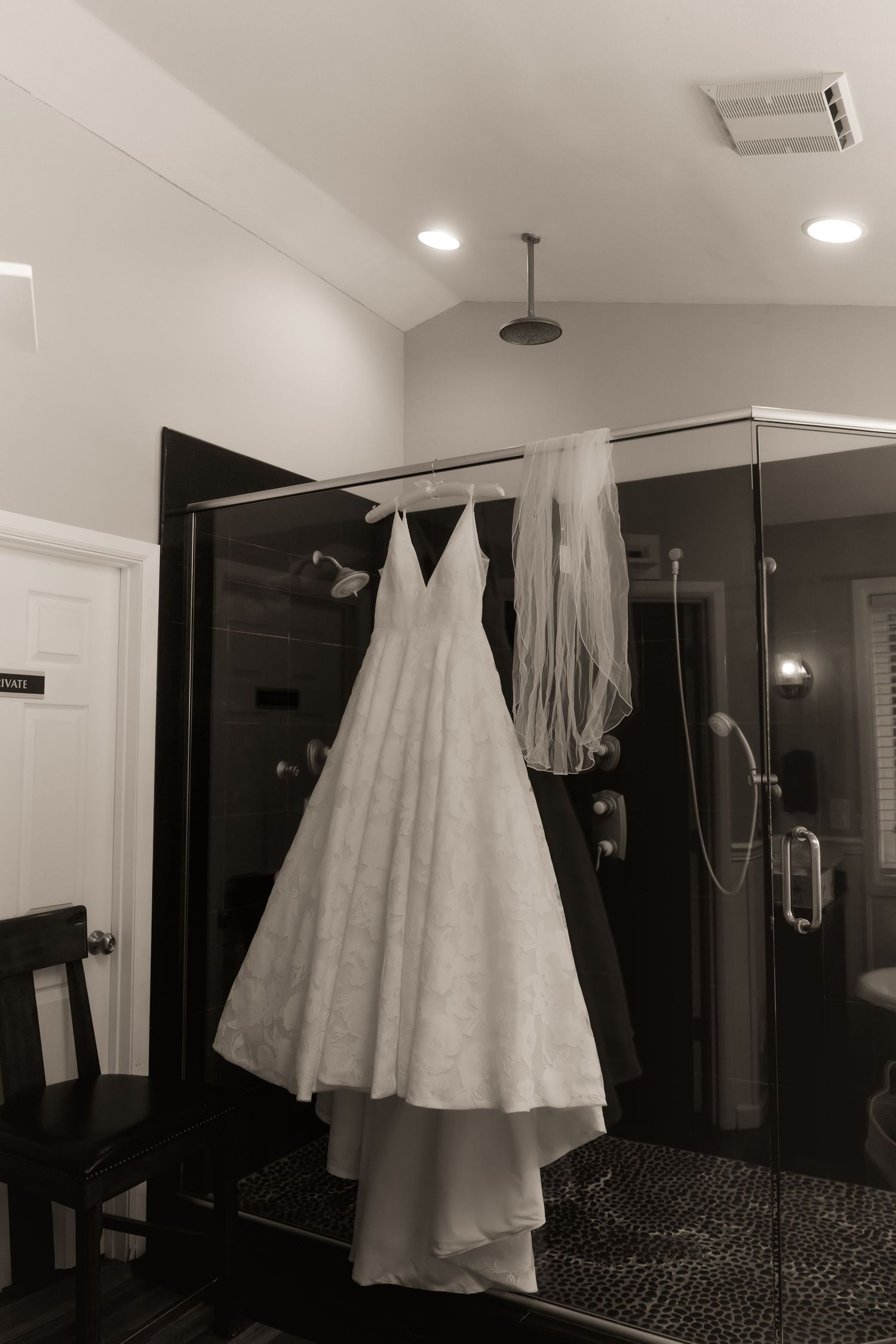 Black and white photo of a wedding dress and veil hanging on a glass shower door in an elegant bathroom.