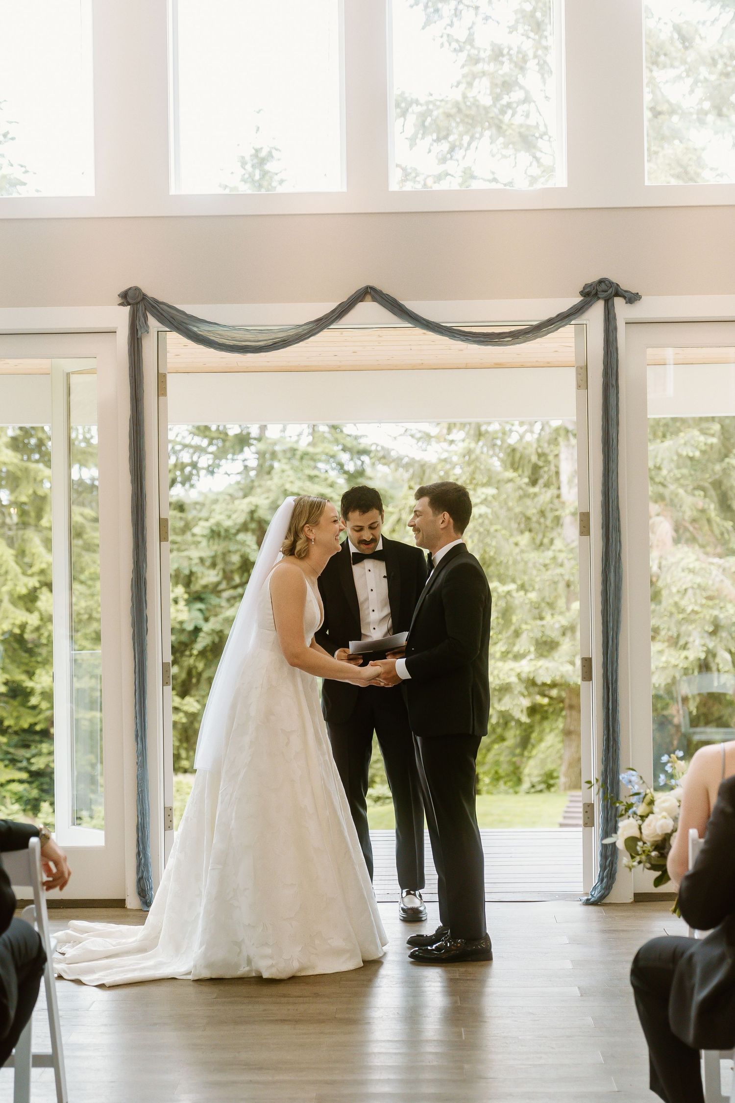 Bride and groom exchange vows at indoor wedding ceremony with large windows and lush greenery backdrop.