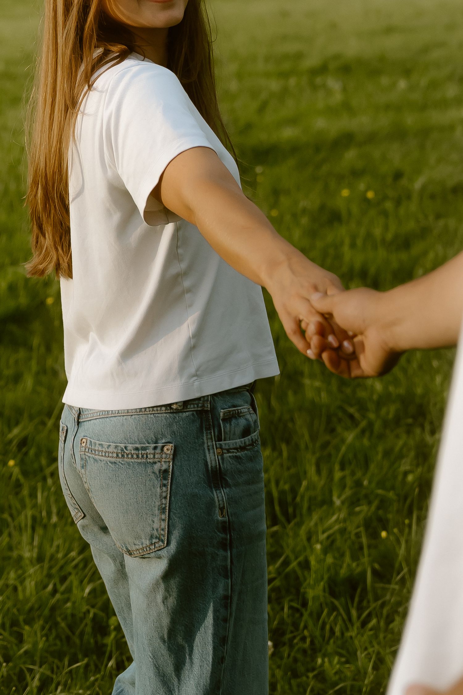 A girl with long hair in a white t-shirt and jeans holds hands with someone in a sunny green field.