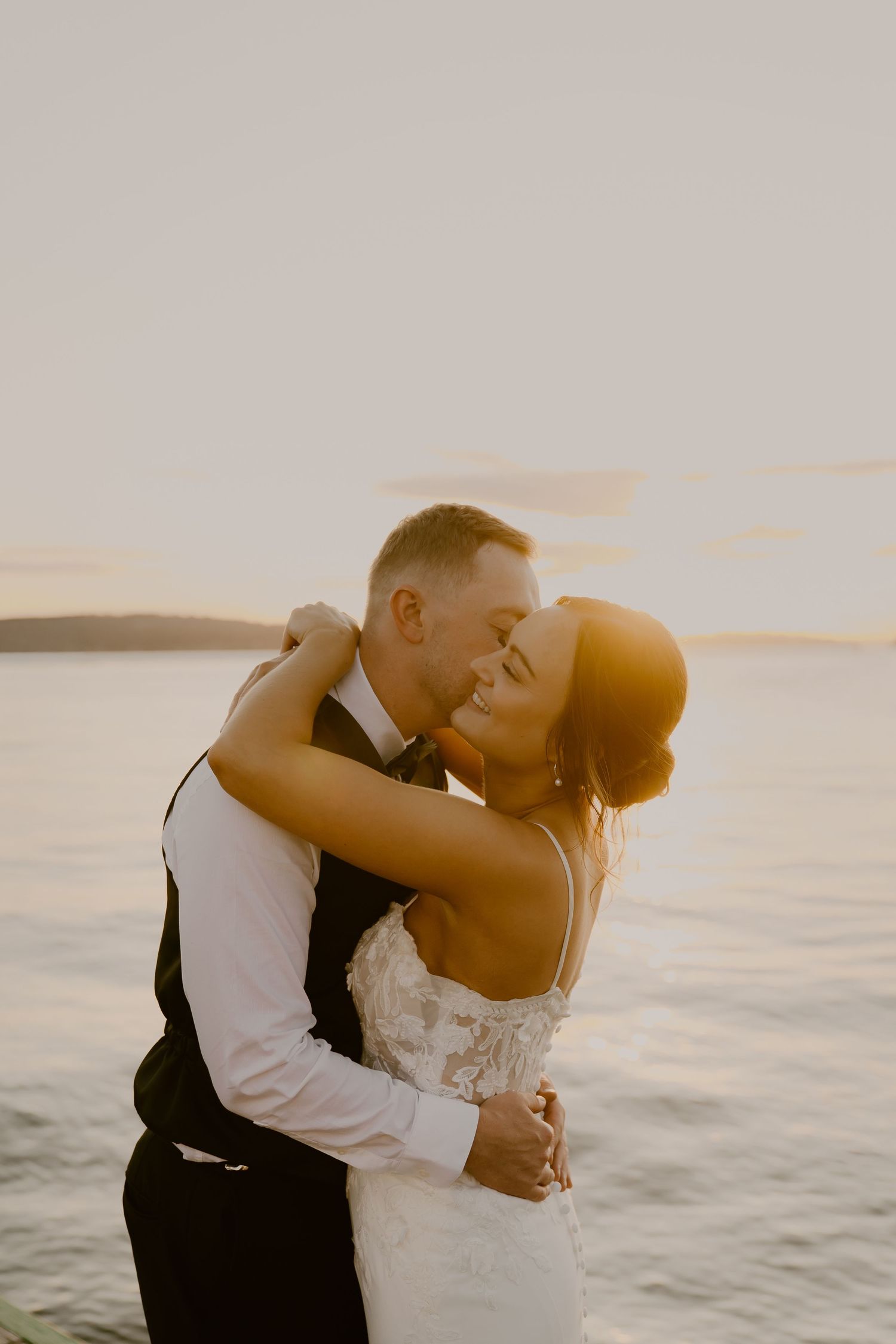 A romantic couple embraces during sunset by the water in wedding attire with golden light illuminating the scene.