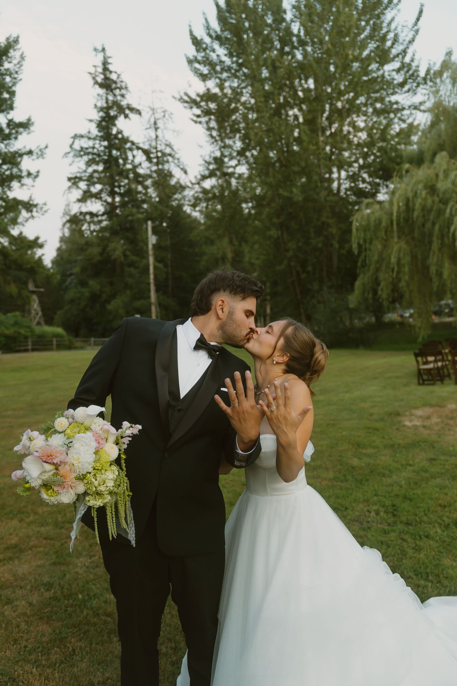 Bride and groom share a kiss outdoors, she holds a pink bouquet in a white gown, he wears a black tuxedo.