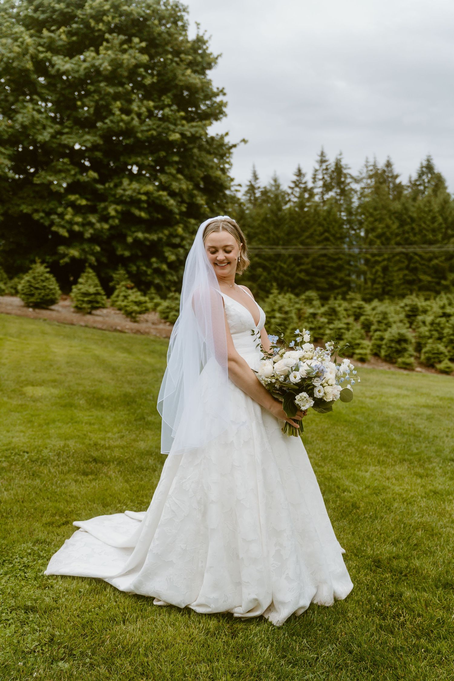 Bride in white lace A-line gown and veil holding bouquet on green lawn with evergreen backdrop.