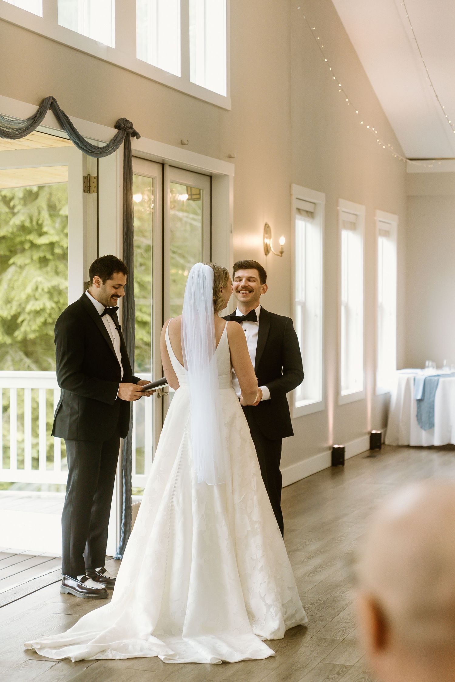Bride and groom exchange vows at indoor wedding ceremony with officiant, bright windows, and elegant white decor.