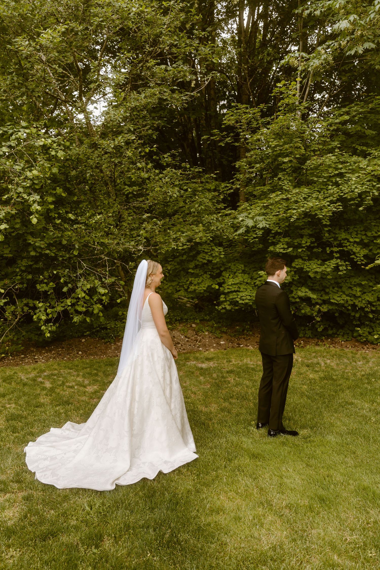 Bride in elegant white gown with veil and groom in black tuxedo share a first look moment on a lush green lawn.