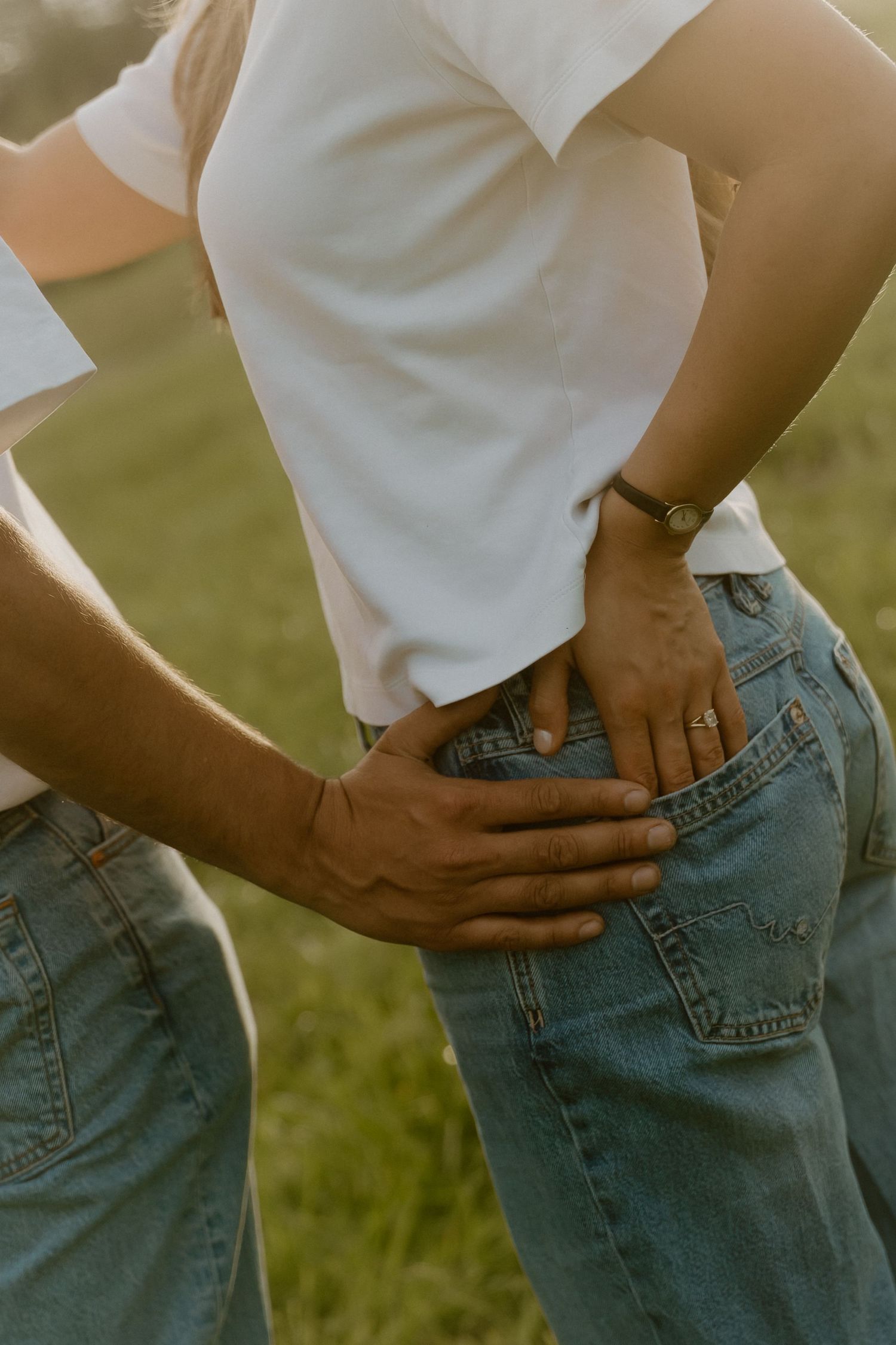 Couple in white tees and blue jeans embrace outdoors at golden hour, hands resting on hips.