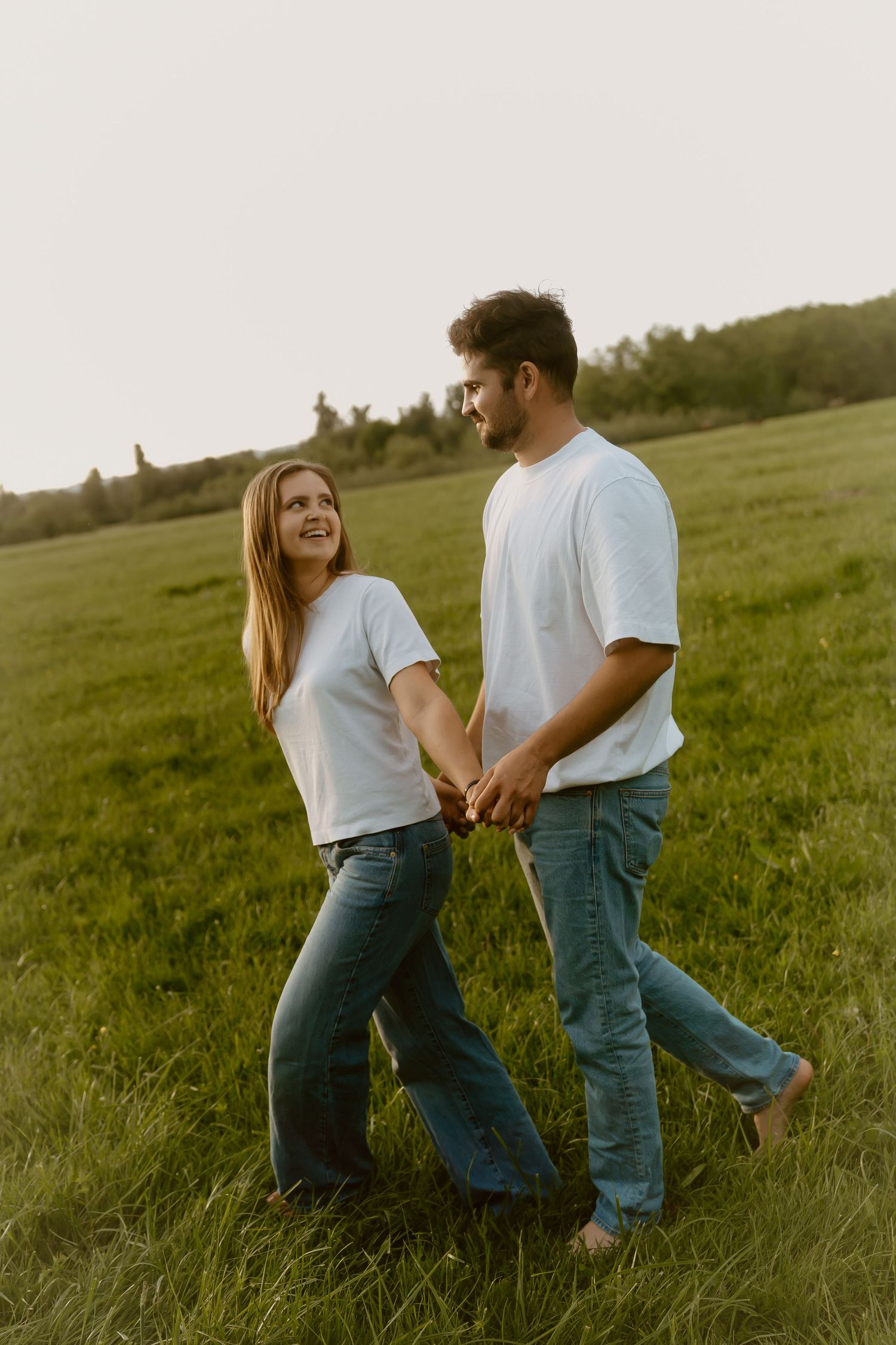 A couple in matching white t-shirts and jeans hold hands while walking through a green field at golden hour.