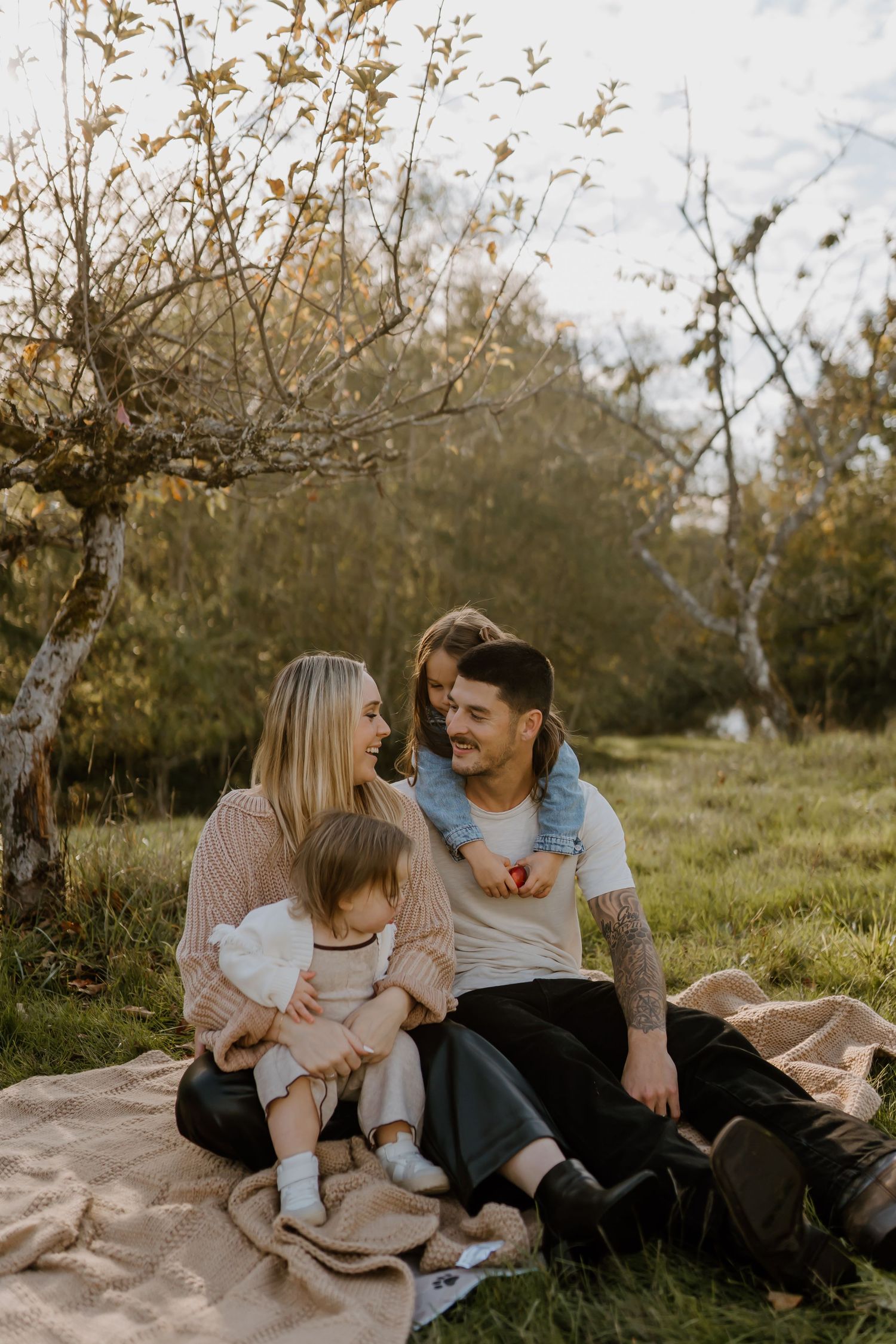 Family sitting on blanket outdoors in autumn park, parents smiling with two young children among golden trees.