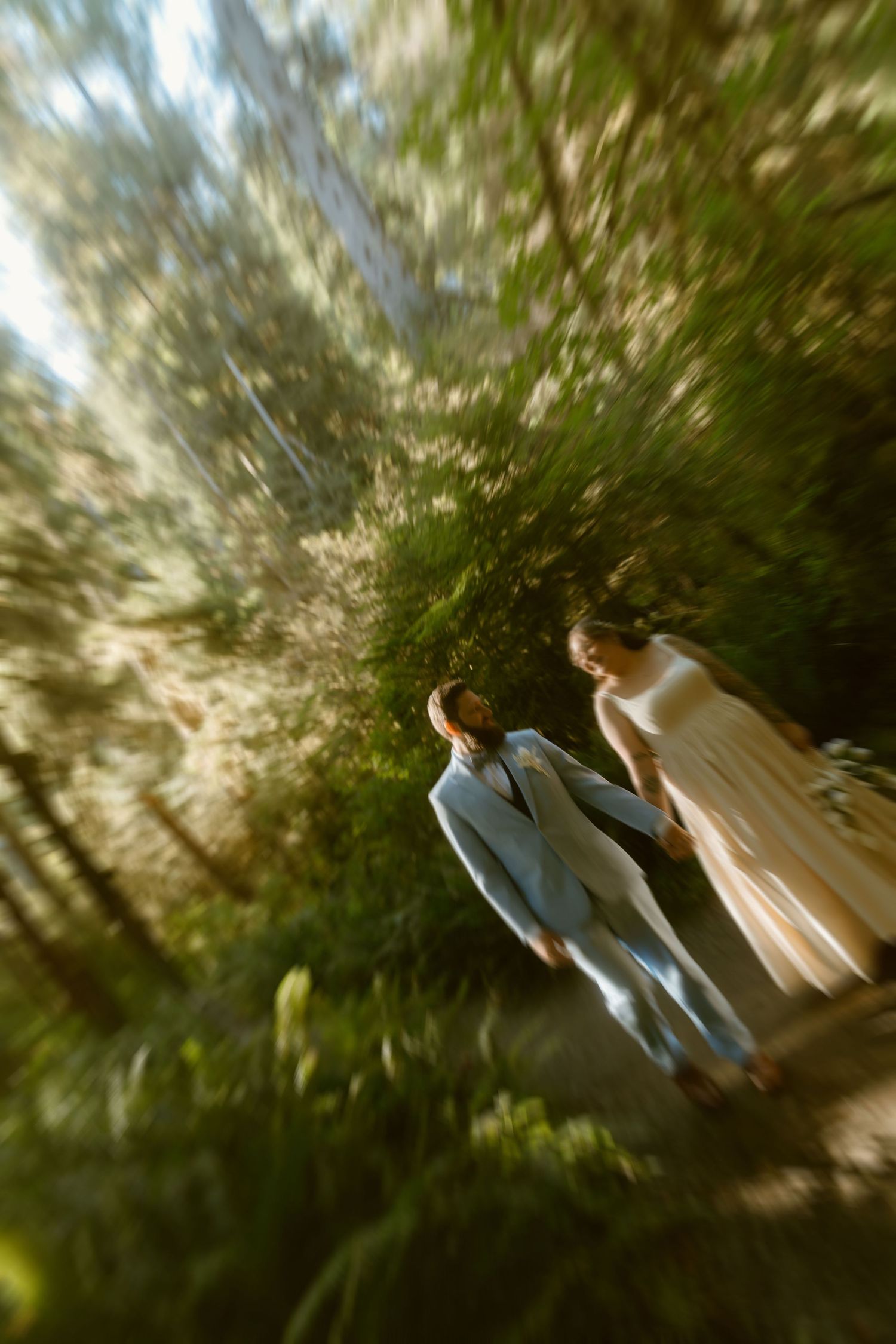 Couple in formal attire walking through lush green forest with dreamy zoom blur effect.