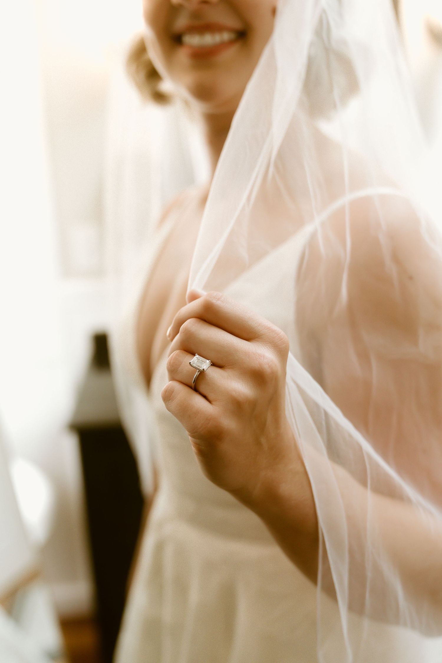 Smiling bride holding her wedding veil, showcasing a stunning diamond engagement ring on her finger.