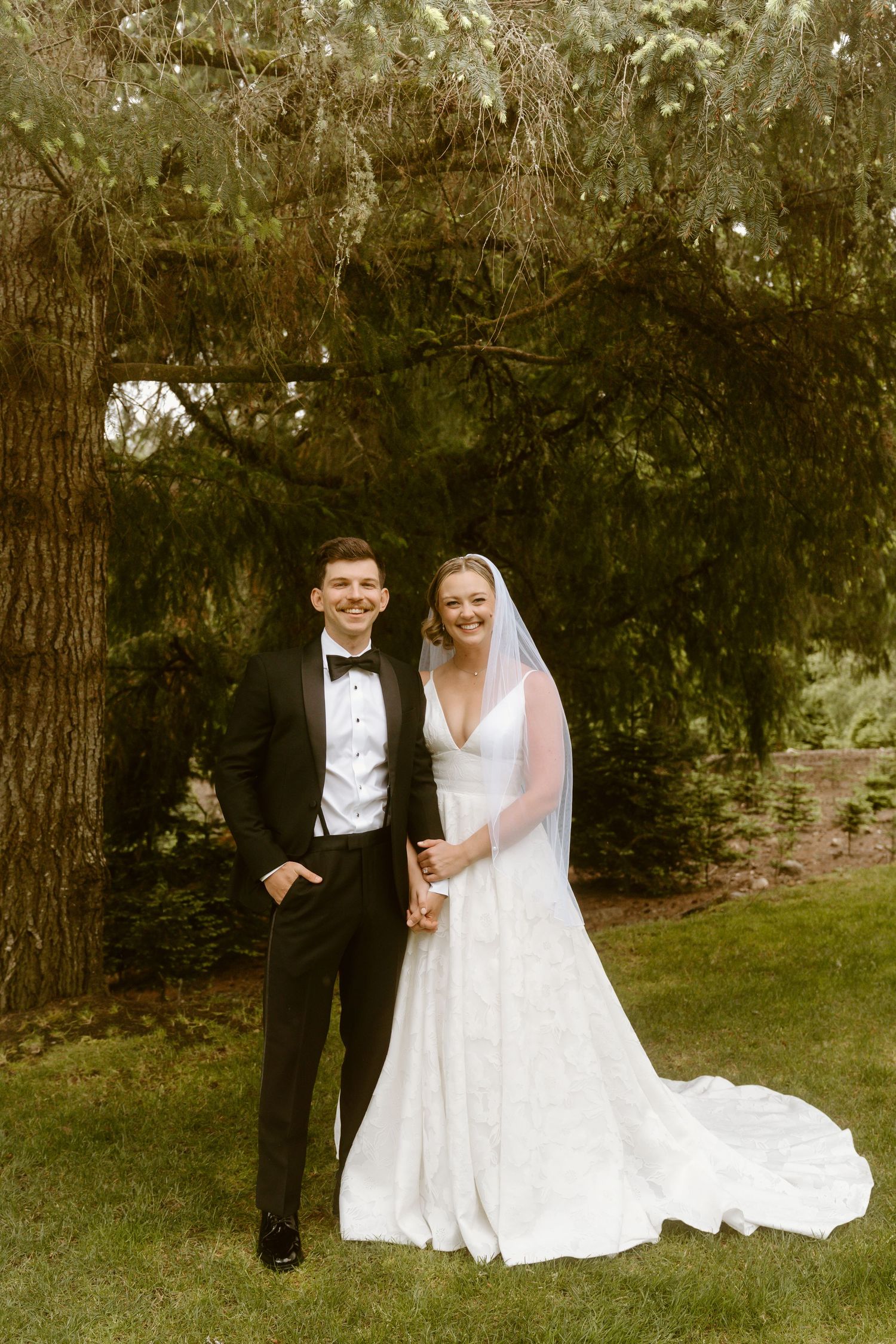 Bride and groom posing together under lush green tree canopy on their wedding day.