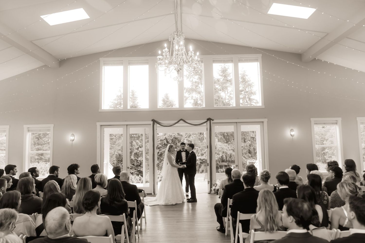 Black and white photo of a wedding ceremony in an elegant chapel with chandelier and large windows.