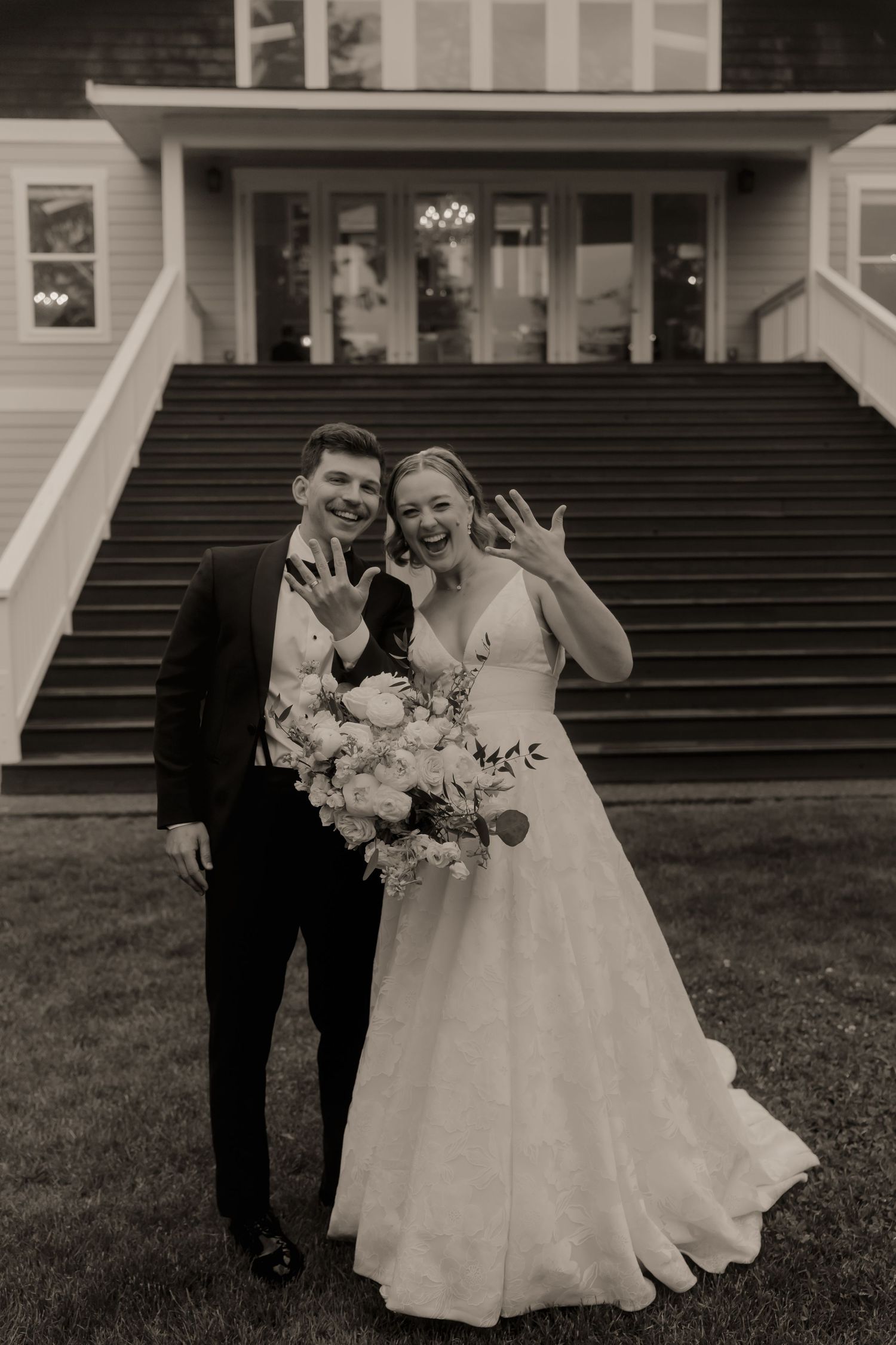 Newlyweds smile and show off wedding rings in black and white photo outside venue with grand staircase.