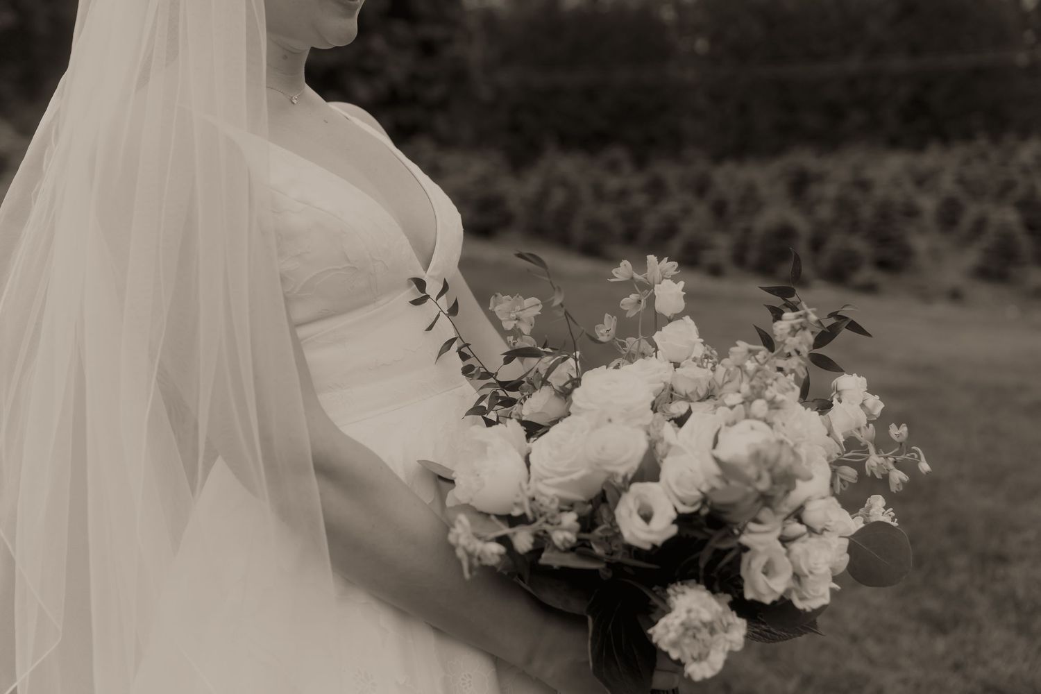Black and white photo of a bride in a strapless gown and veil holding a lush floral bouquet outdoors.