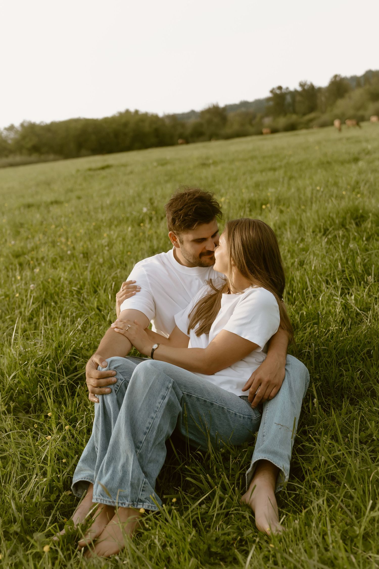 Couple in white t-shirts and jeans sitting barefoot on green grass field, embracing romantically at sunset.