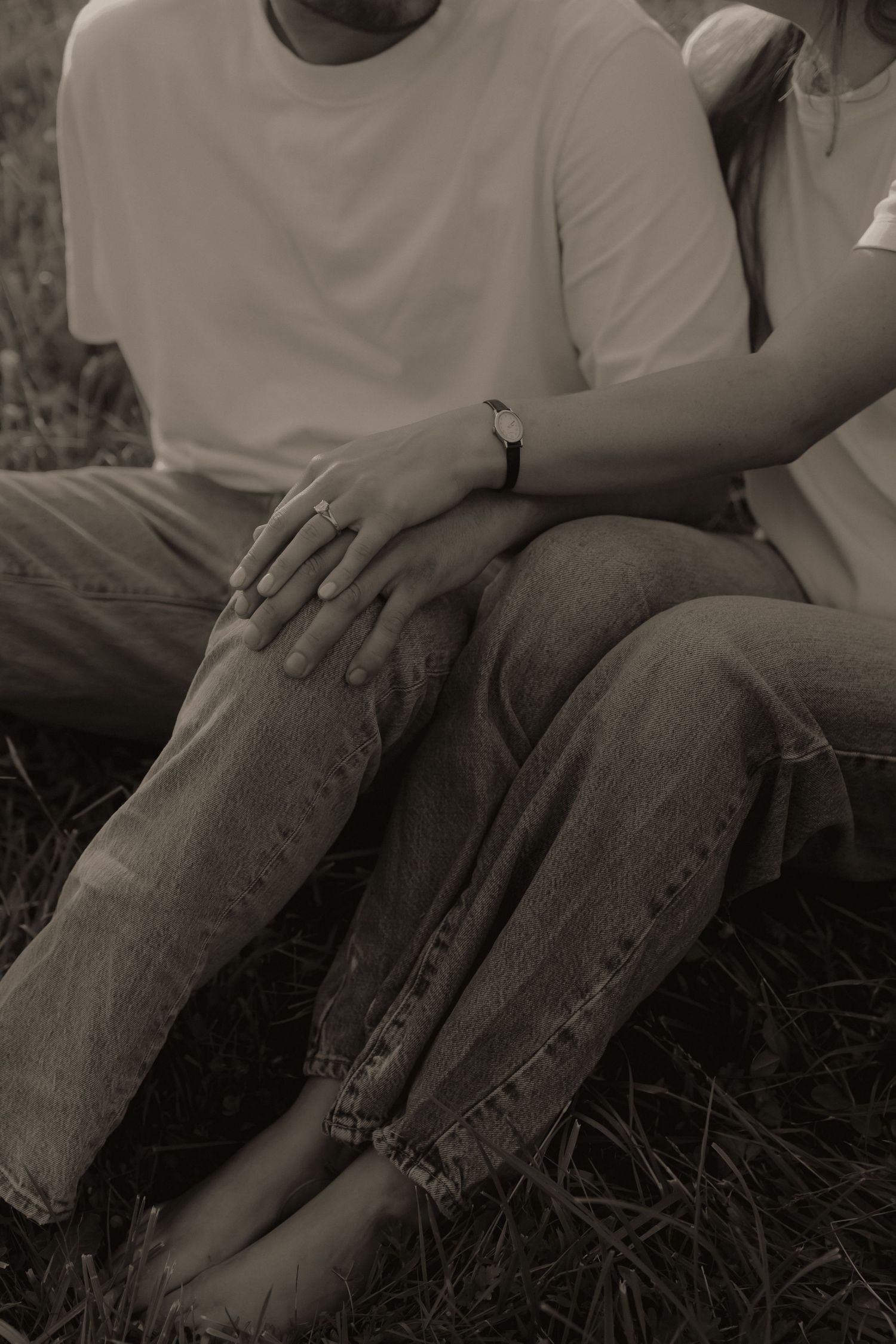 Sepia-toned photo of a couple sitting on grass, hands intertwined, wearing jeans and casual tops.