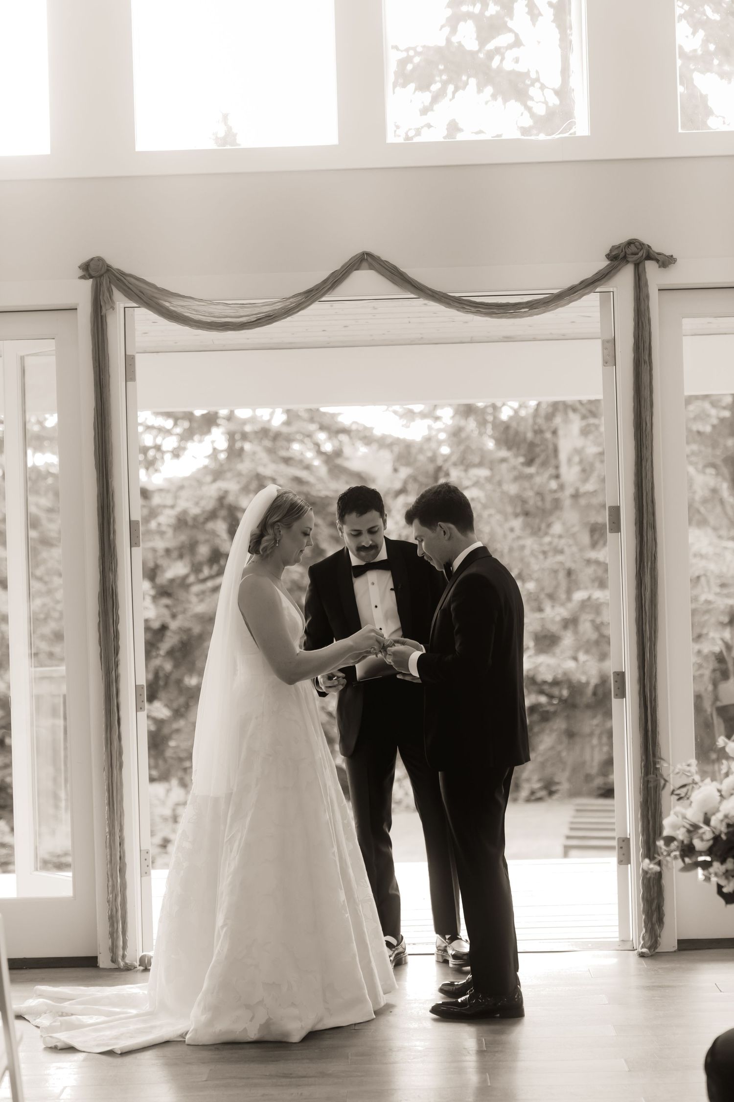 Black and white photo of bride and groom exchanging rings with officiant at wedding altar.