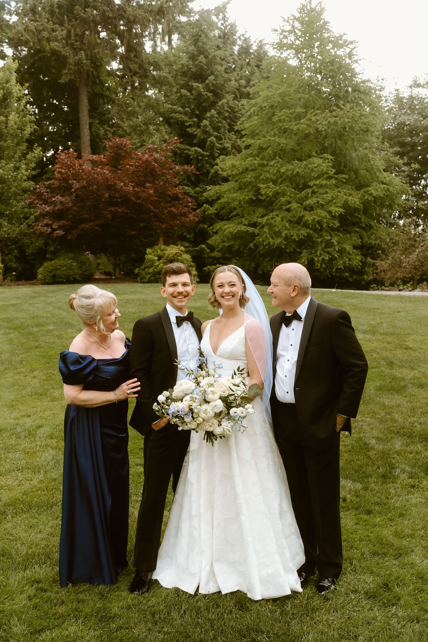 Wedding couple poses with parents in formal attire on green lawn with tall trees in background.