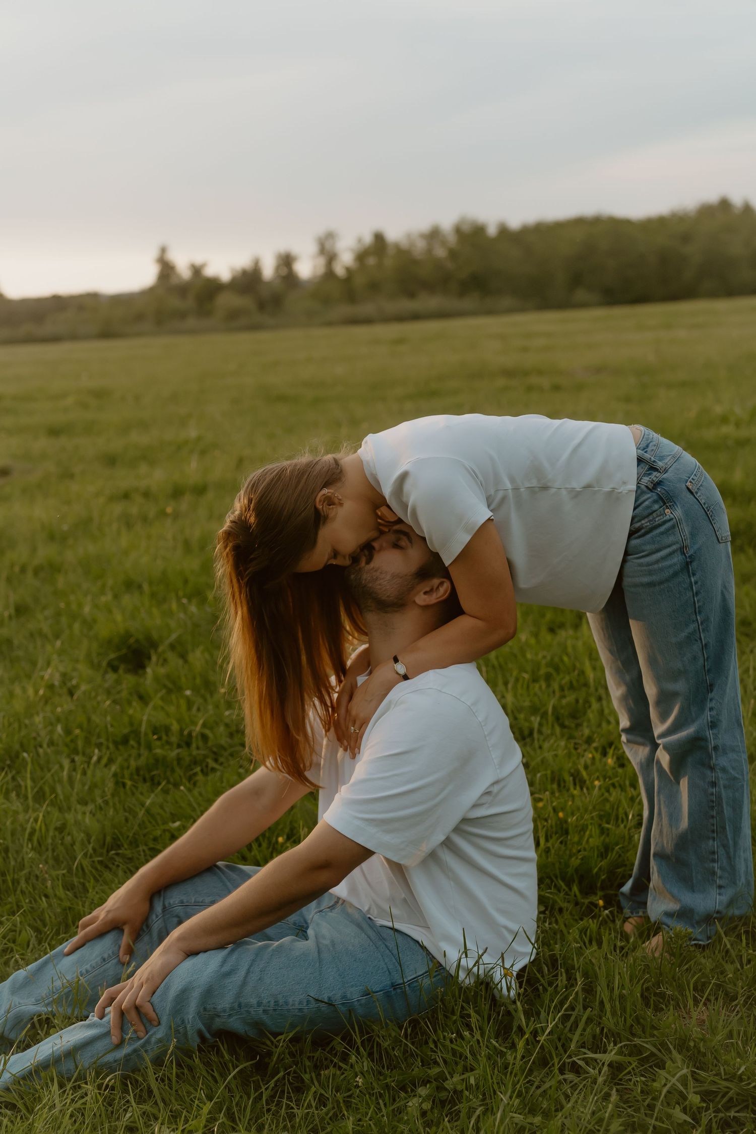 Romantic outdoor moment as woman leans down to kiss man sitting on grass in a green field.