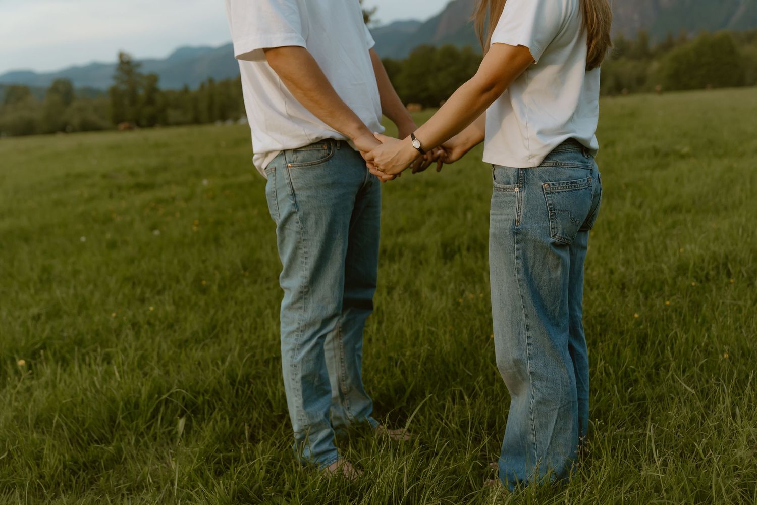 Couple holding hands in a green field with mountains, wearing white shirts and blue jeans.