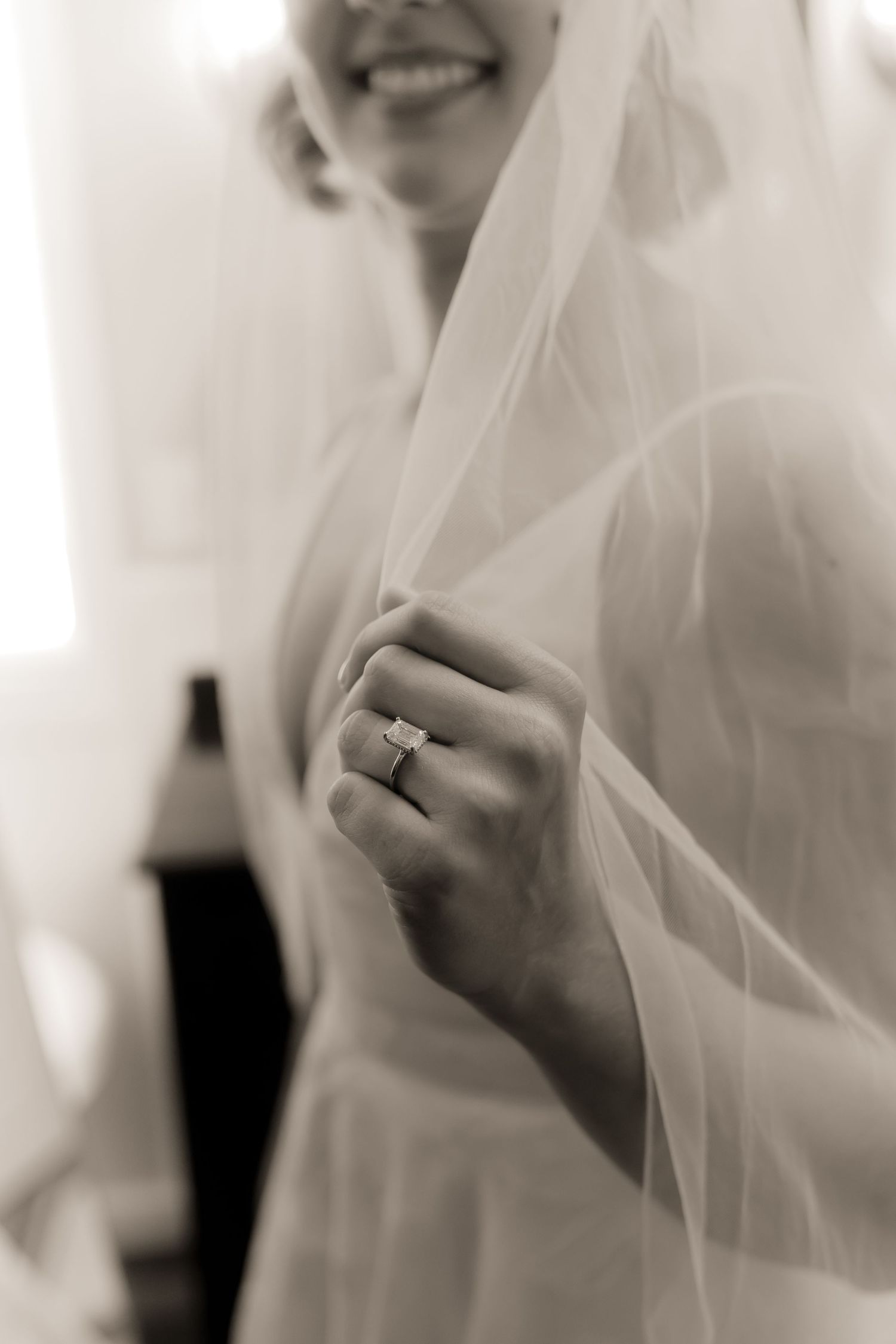 Black and white photo of a bride holding her veil, showcasing a stunning diamond engagement ring on her finger.