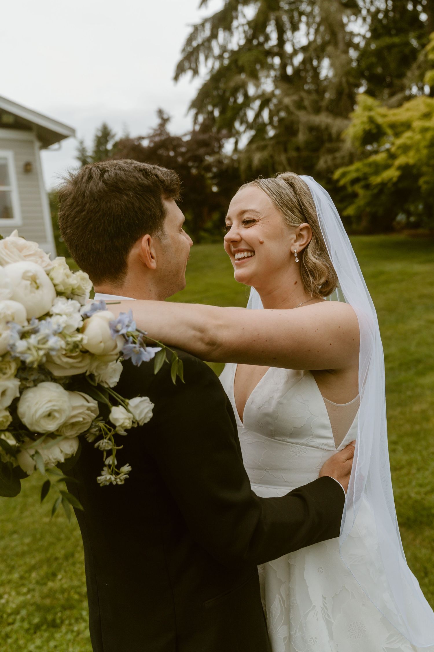 Laughing bride in white gown embraces groom in black tuxedo during outdoor wedding portraits.